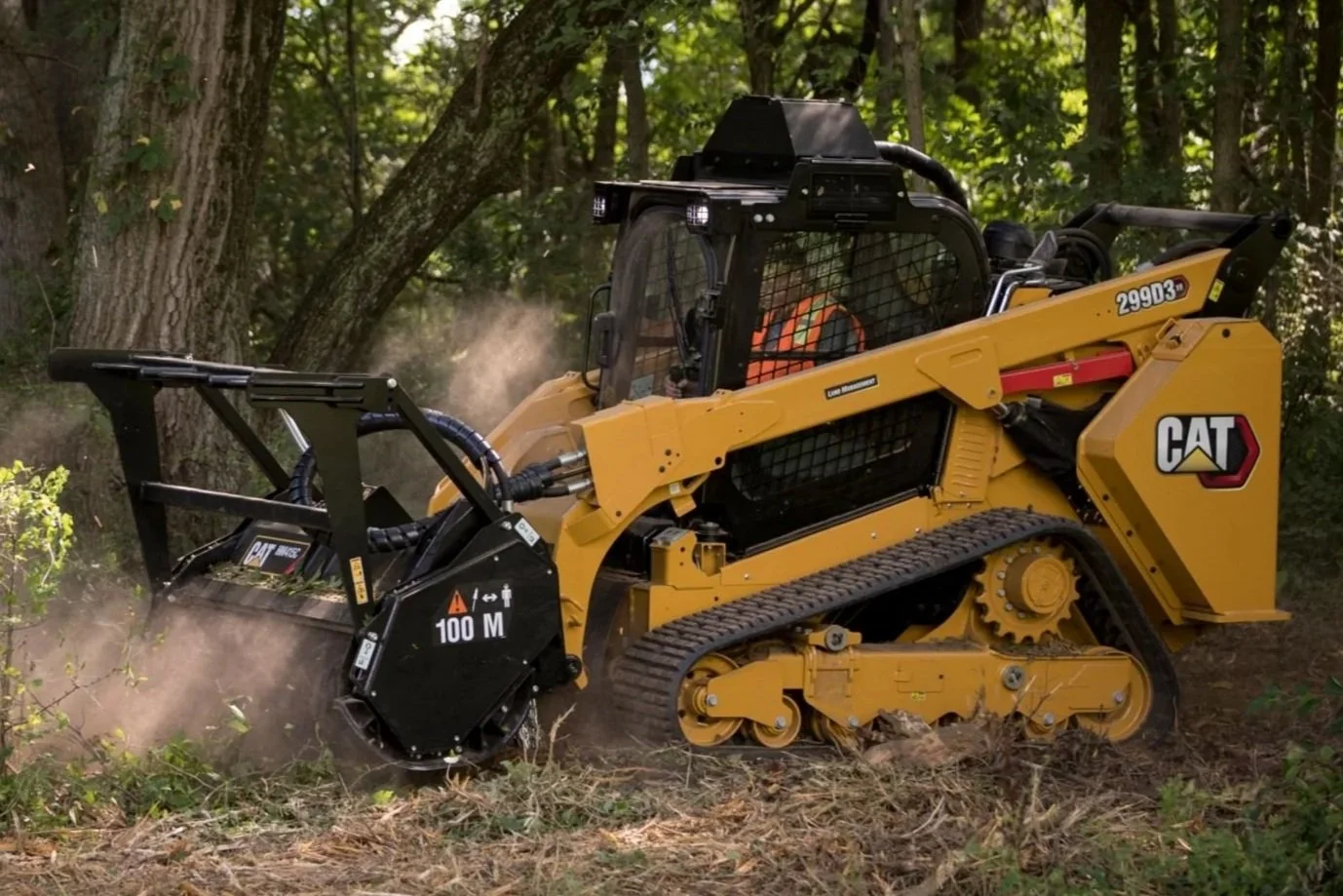A yellow compact track loader skid steer with a black brush cutter muncher attachment clearing brush in a wooded area. Land clearing, mulching overgrowth and removing brush is it’s specialty.