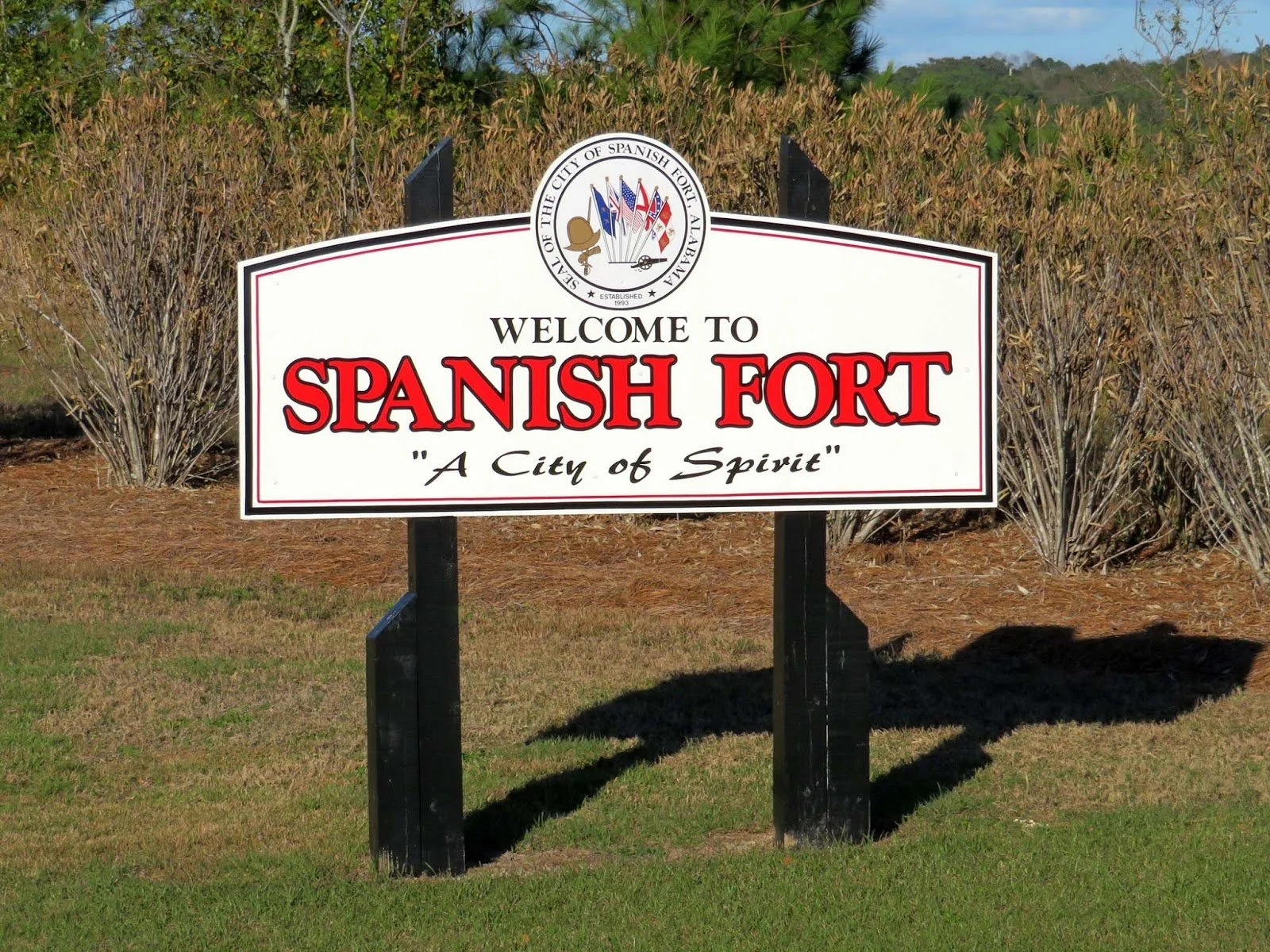 A welcome sign for Spanish Fort, with a city motto 'A City of Spirit', surrounded by shrubbery and grass.