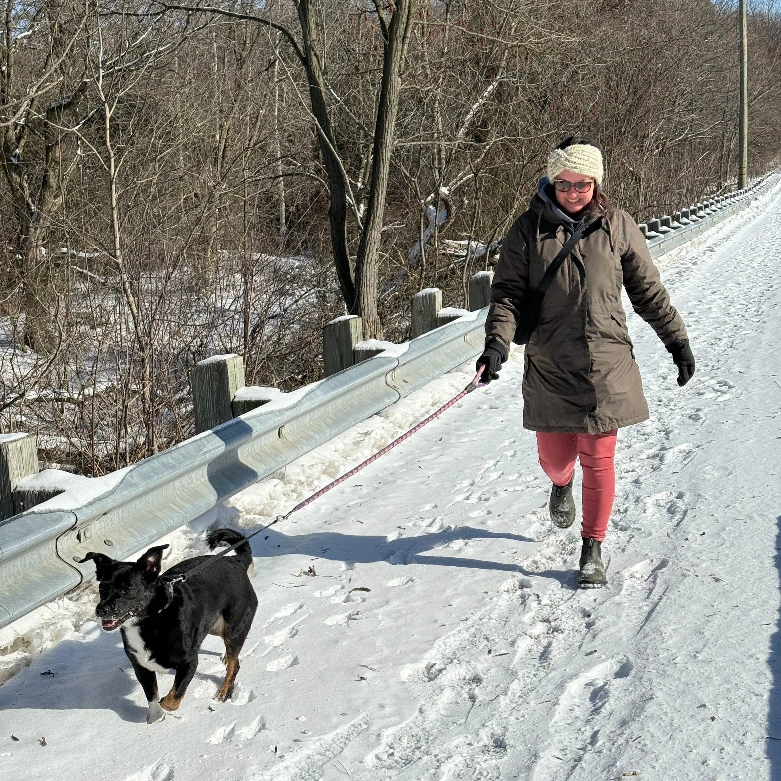 Crystal, who is wearing a long winter coat, pink pants, and a white knitted hat, is walking a small black and tan dog on a snowy path.