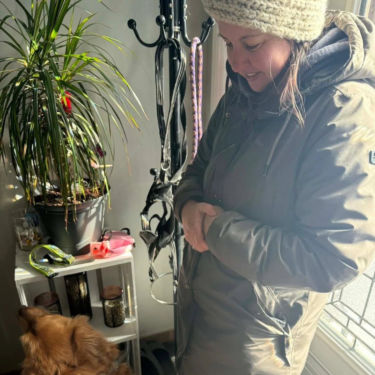 Crystal wearing a coat and hat, smiling inside her house beside a dog, with a coat rack with dog leads hanging from it in the background.