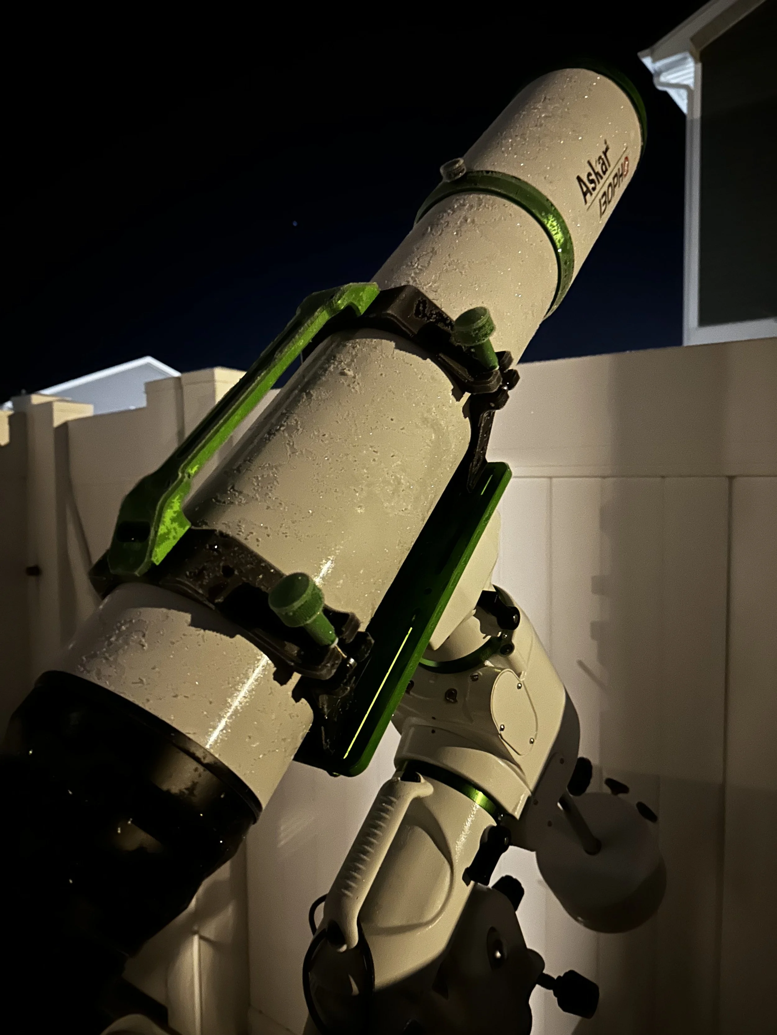 A close-up view of a telescope labeled 'Askara' and 'BOOH,' with water droplets on its surface, pointing upwards against a dark sky at night.