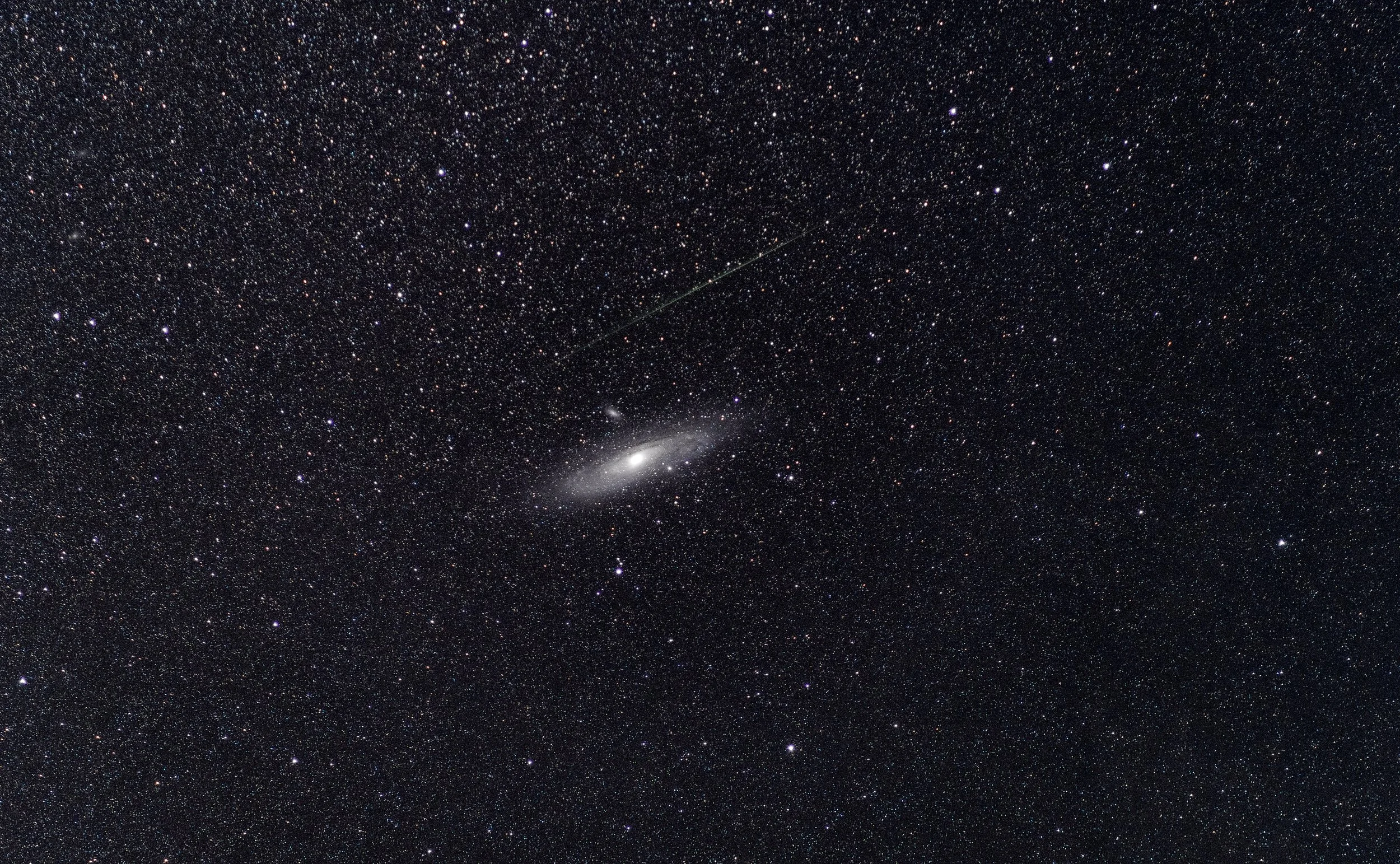 A photograph of a star-filled night sky showing countless stars, a galaxy at the bottom center, and a faint shooting star or meteor streaking diagonally across the sky.