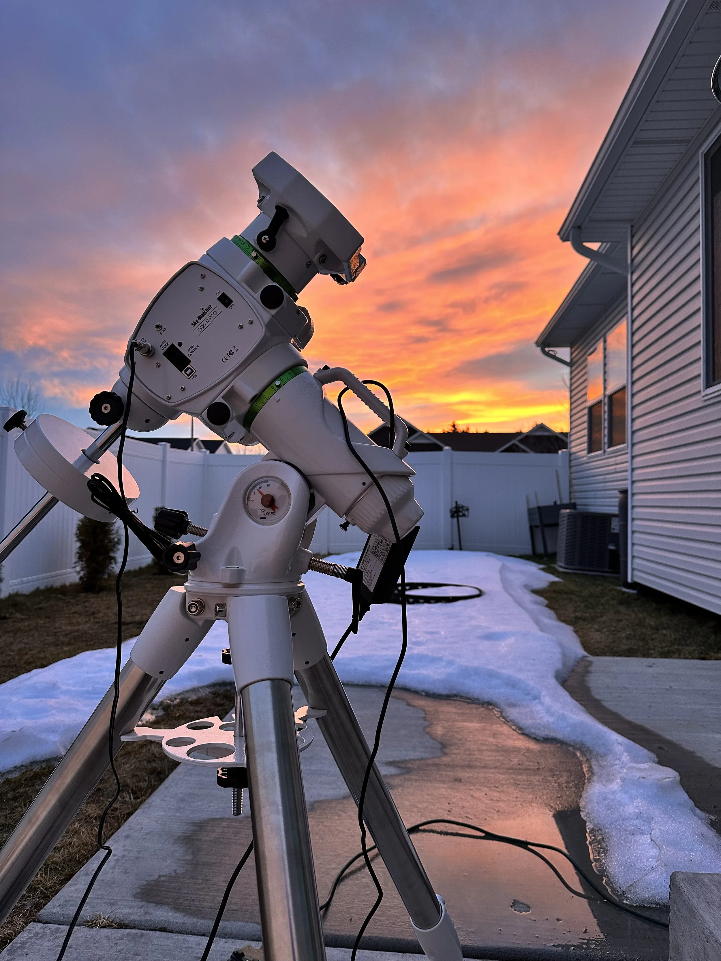 A white telescope set up outside during sunset with a colorful sky, adjacent to a house with vinyl siding and a concrete sidewalk with patches of snow.