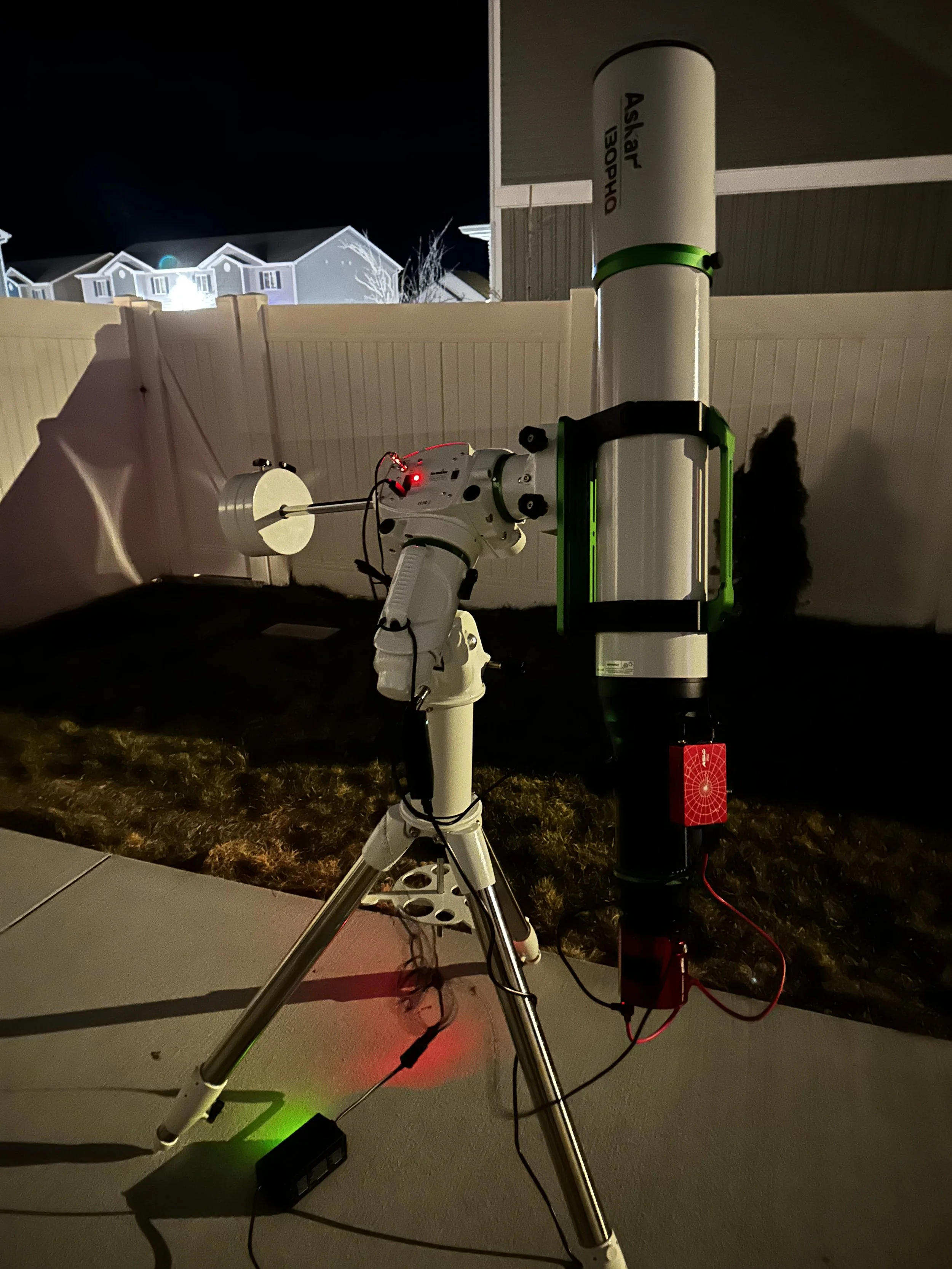 A telescope mounted on a tripod pointed at the night sky, with a green light illuminating part of the telescope, set up outdoors at night with a white fence and houses in the background.