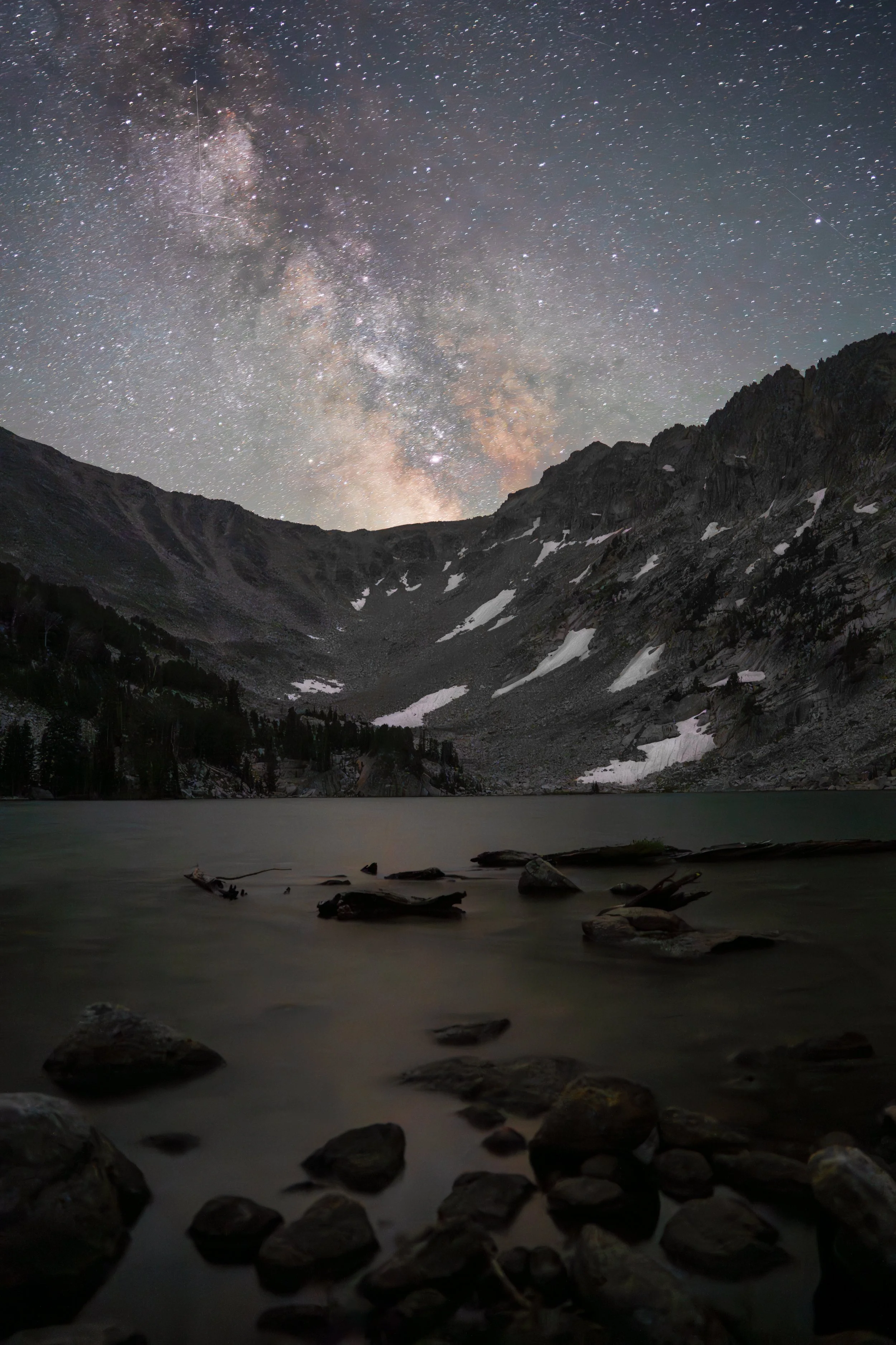 Nighttime view of a star-filled sky above a mountain landscape with snow patches, overlooking a calm lake with rocks in the foreground.