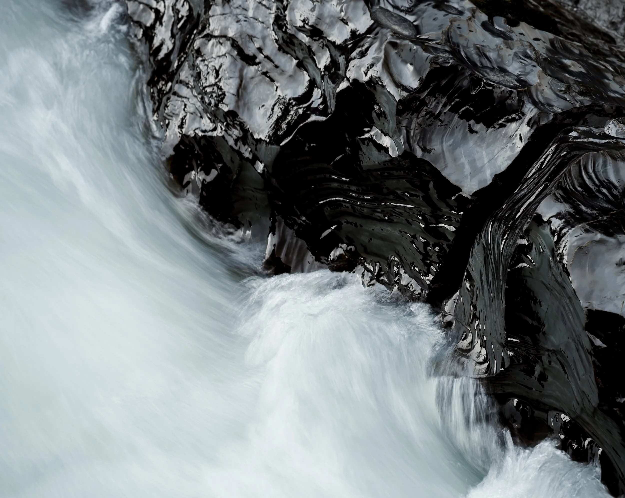 Close-up of a flowing icy river, with dark, jagged rocks on one side and white, frothy water on the other.