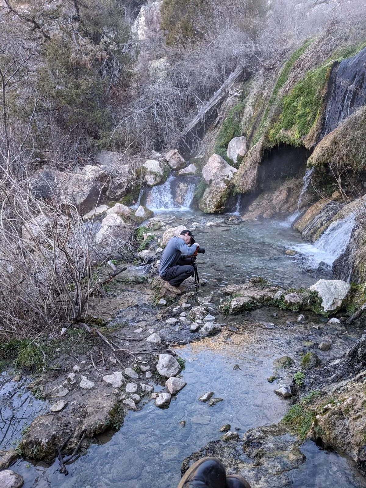 A person is crouched down by a creek, adjusting a camera on a tripod, surrounded by rocks, trees, and a small waterfall in a natural outdoor setting.