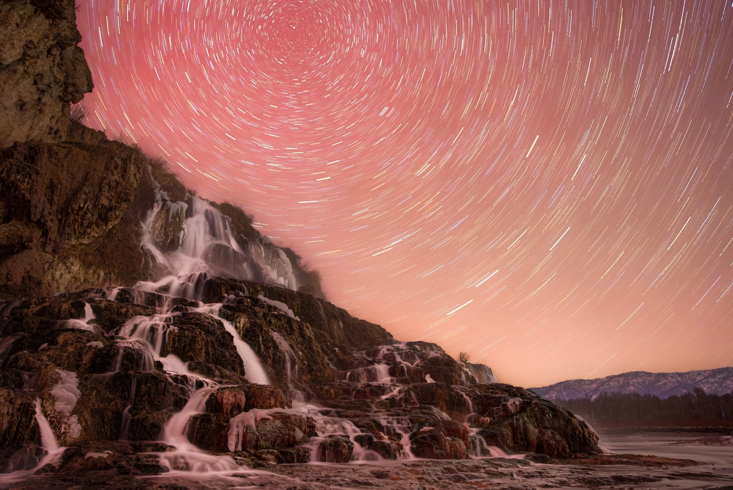 Star trails over a waterfall and rocky landscape at night with a pinkish sky.