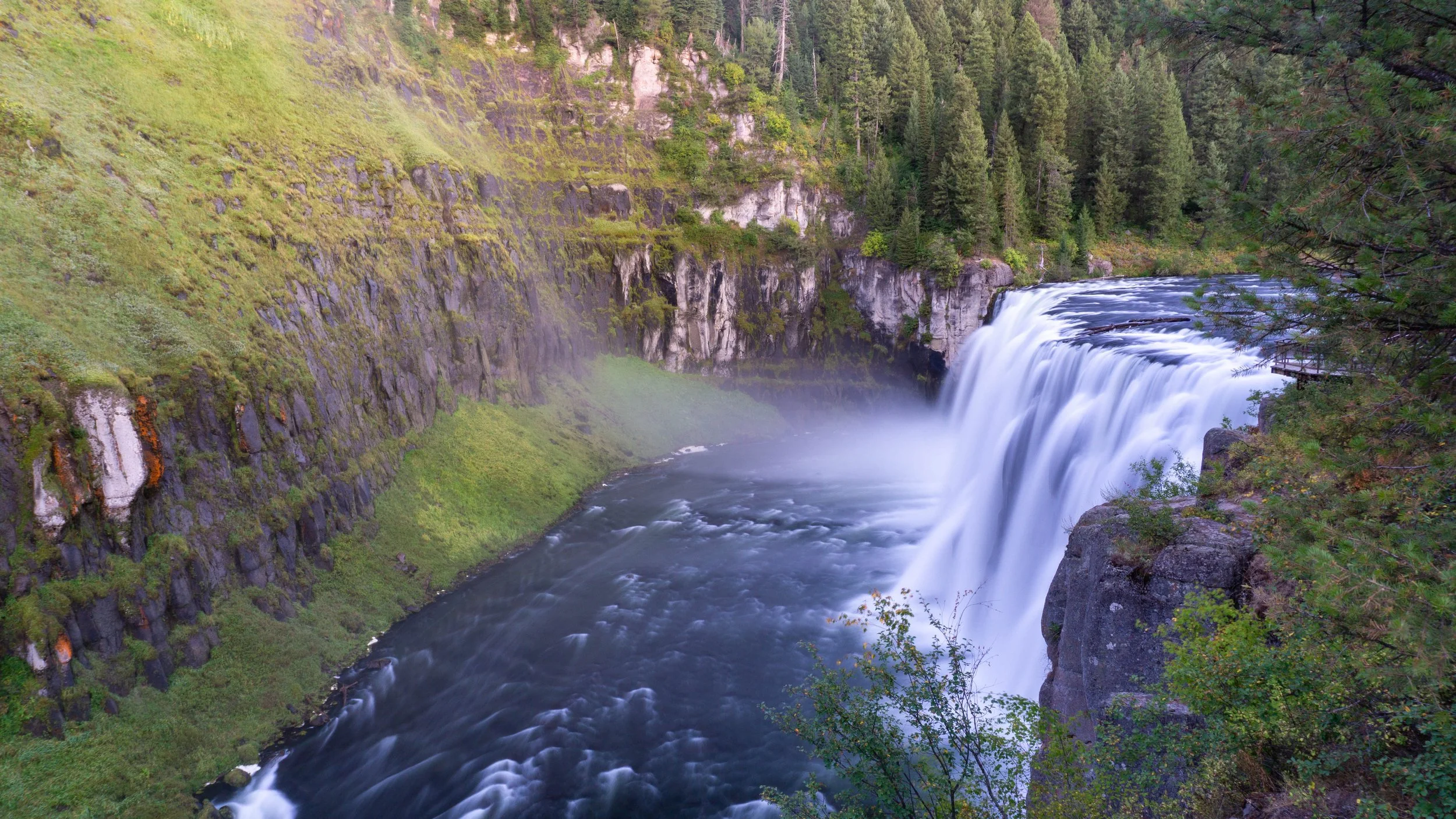 A scenic view of a waterfall flowing into a river, surrounded by lush green trees and rocky cliffs.