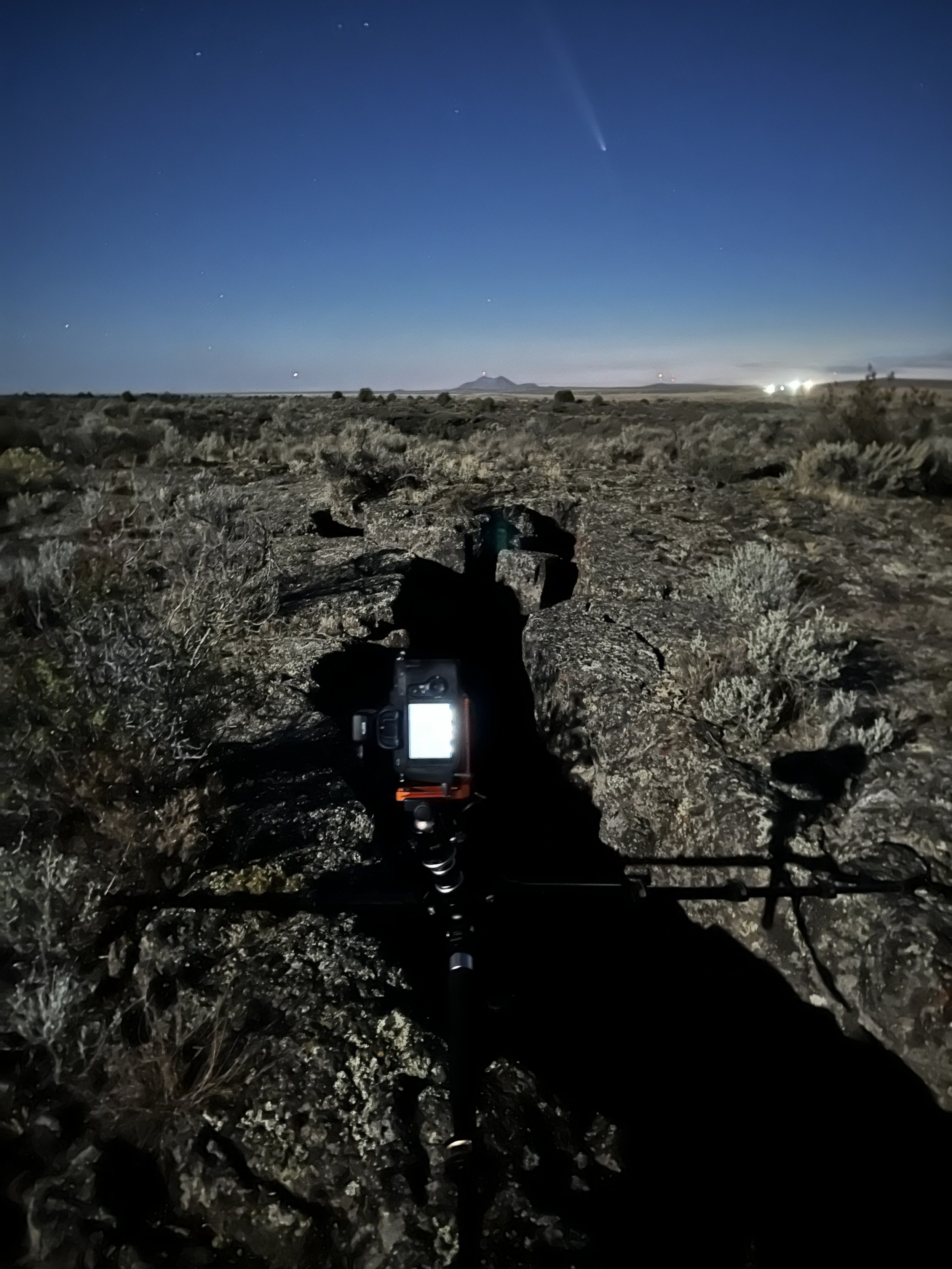 Nighttime desert landscape with rocky terrain and sparse vegetation, a camera mounted on a tripod in the foreground, distant mountains, a clear dark sky with visible stars and a bright comets or shooting star.