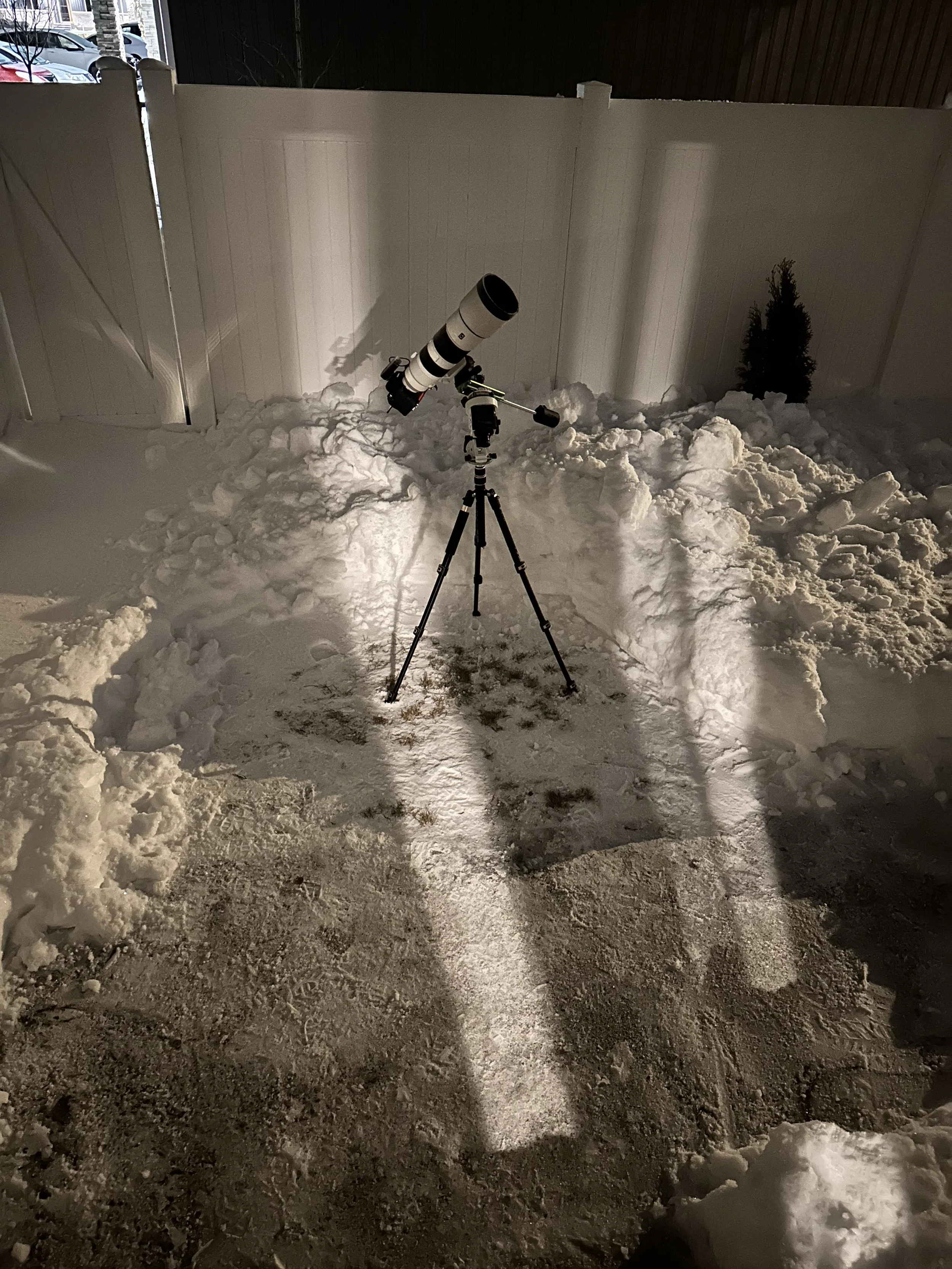A telescope on a tripod set up in a snowy outdoor area at night, with a white fence and small pine tree in the background.