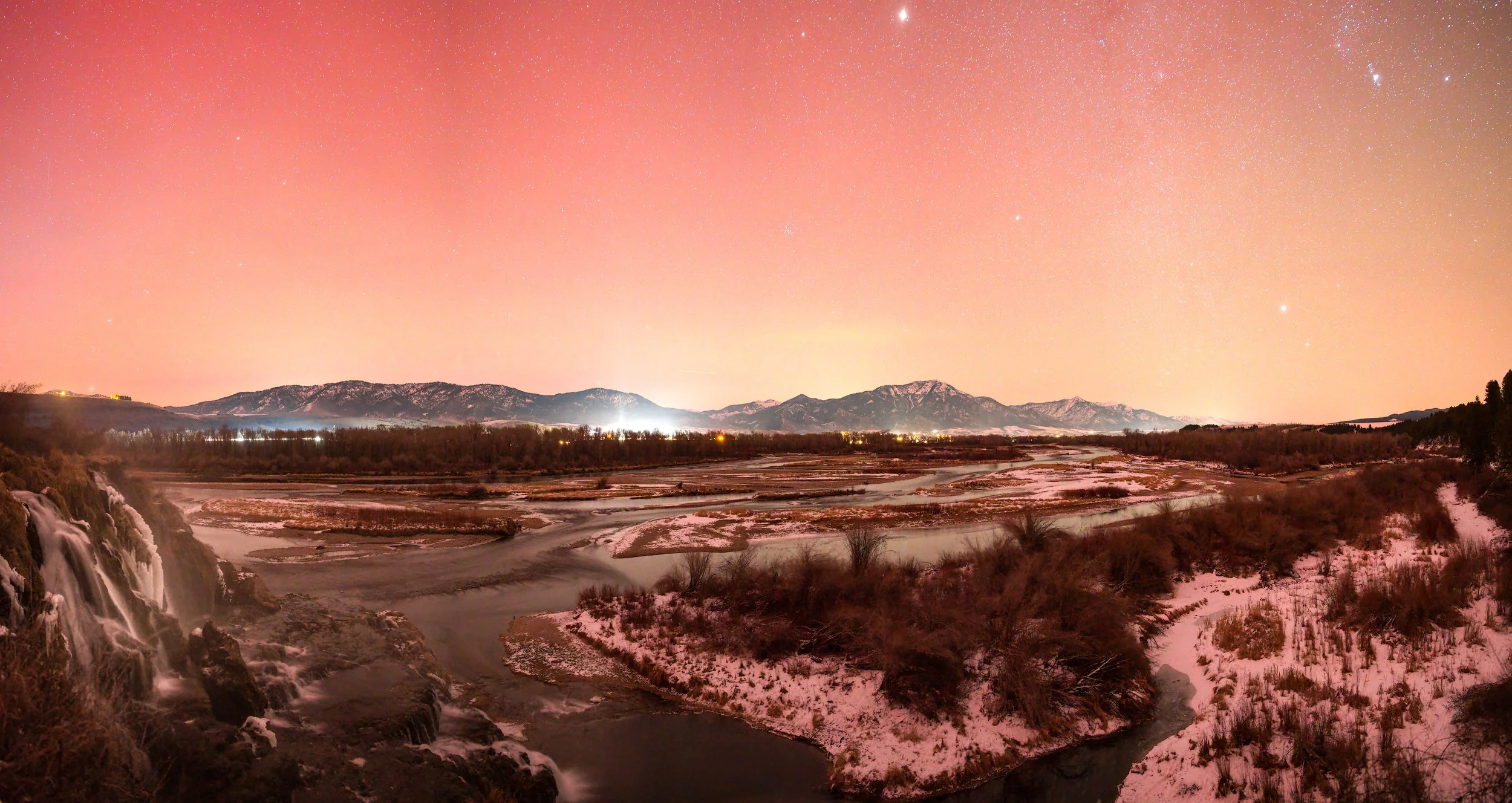 Nighttime landscape with a waterfall to the left, a river flowing through a snowy area, mountains in the distance, and a starry sky with a pinkish hue.