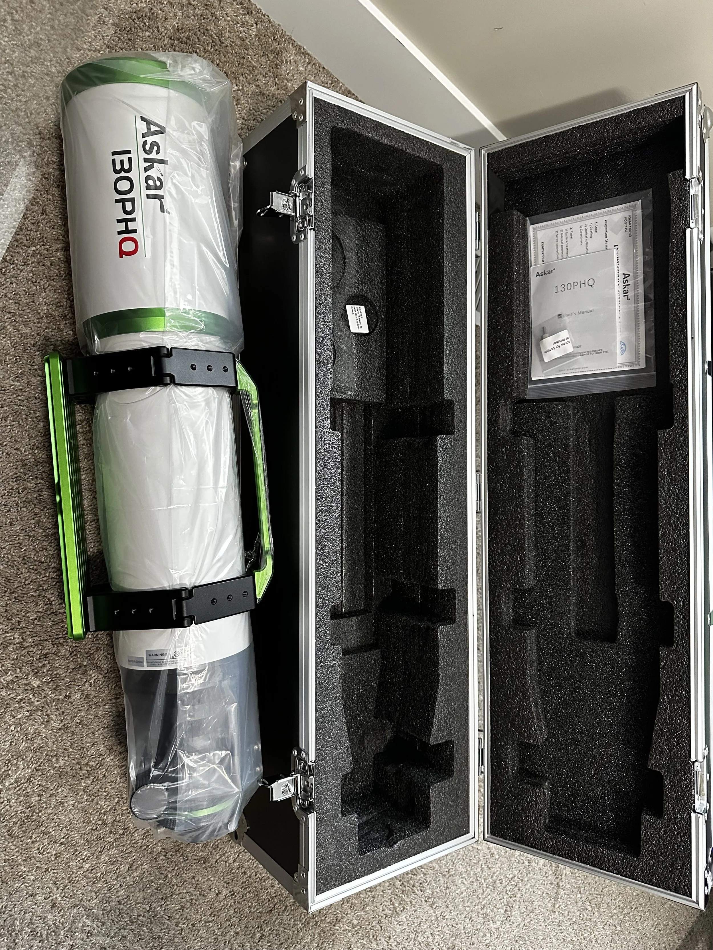 Open black and silver equipment case with foam padding inside, placed on a beige carpeted floor. Behind the case, a long cylindrical object wrapped in plastic labeled 'Ask' and '130PHQ' with a warning label at the bottom, and a handle is attached to the wrapped object.