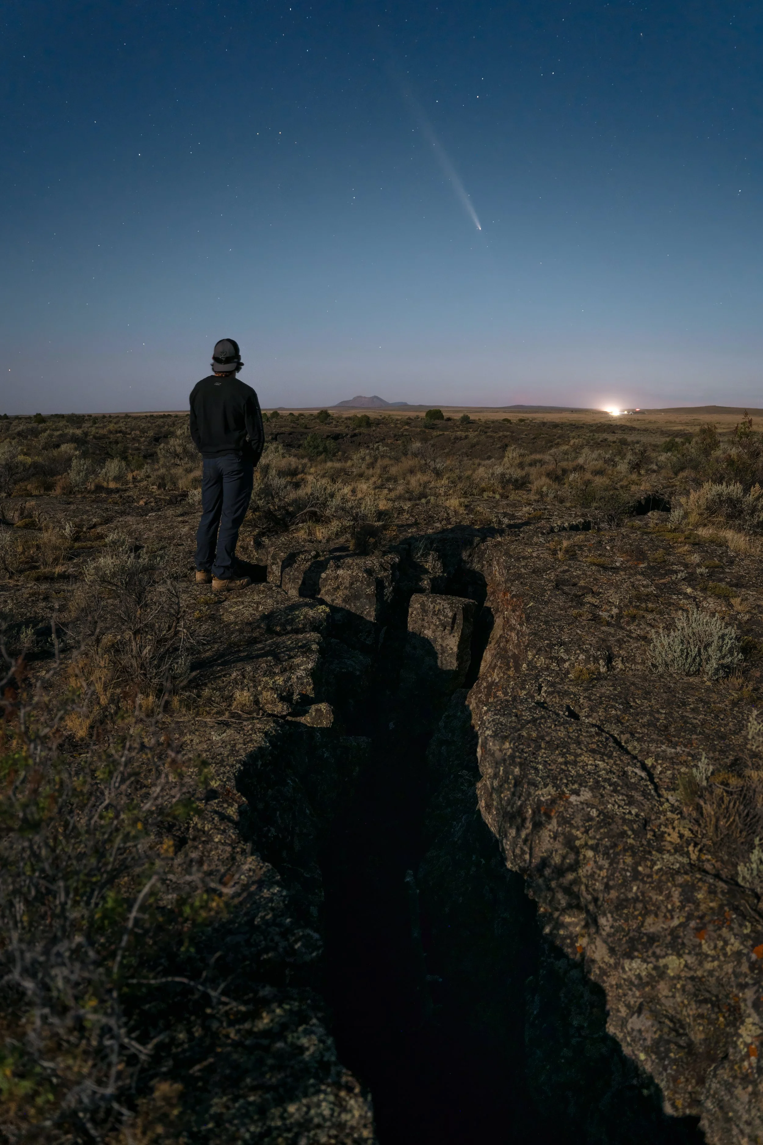 Person standing on rocky terrain at night, observing a starry sky with a comet visible.