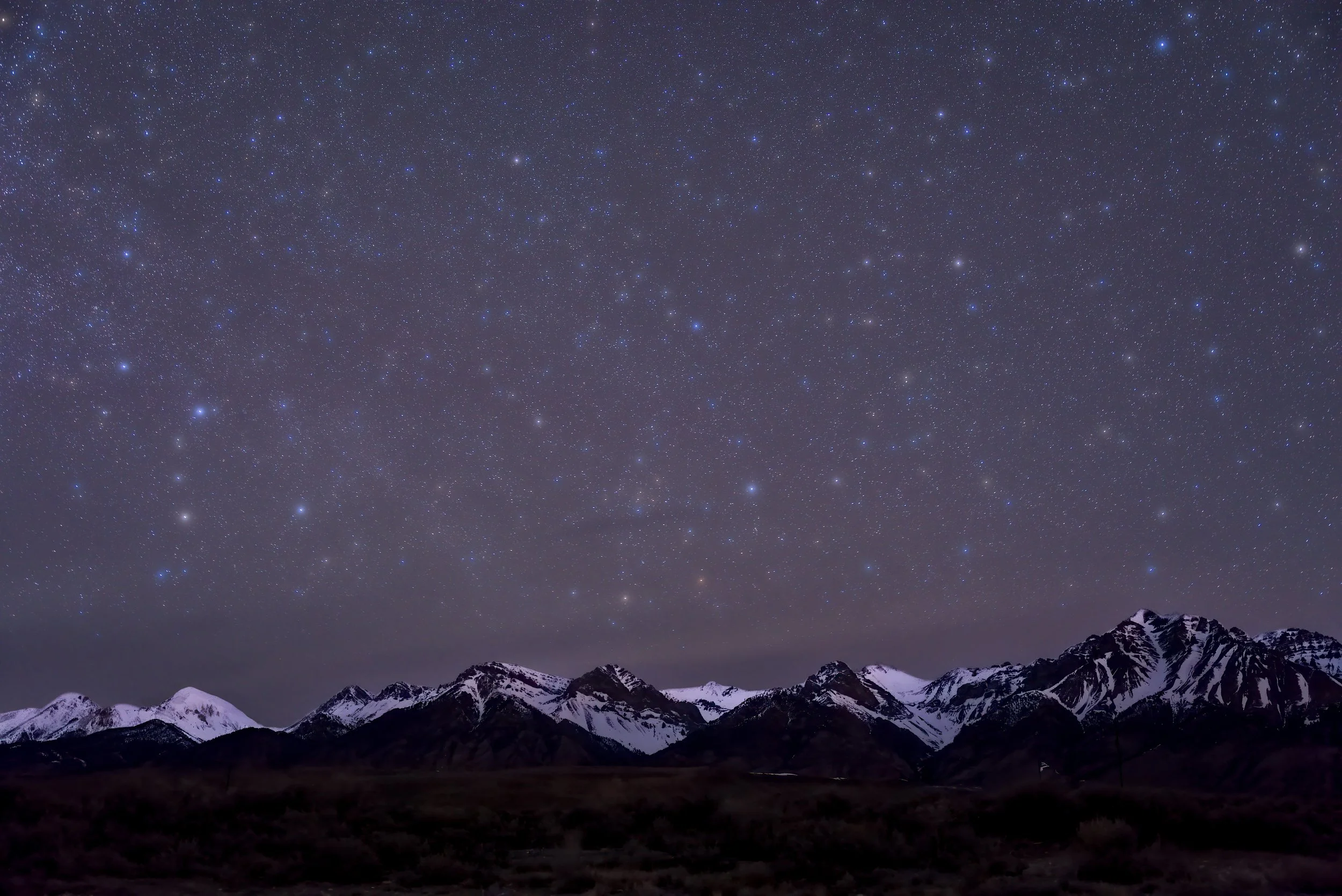 Night sky filled with stars over snow-capped mountains and dark terrain.