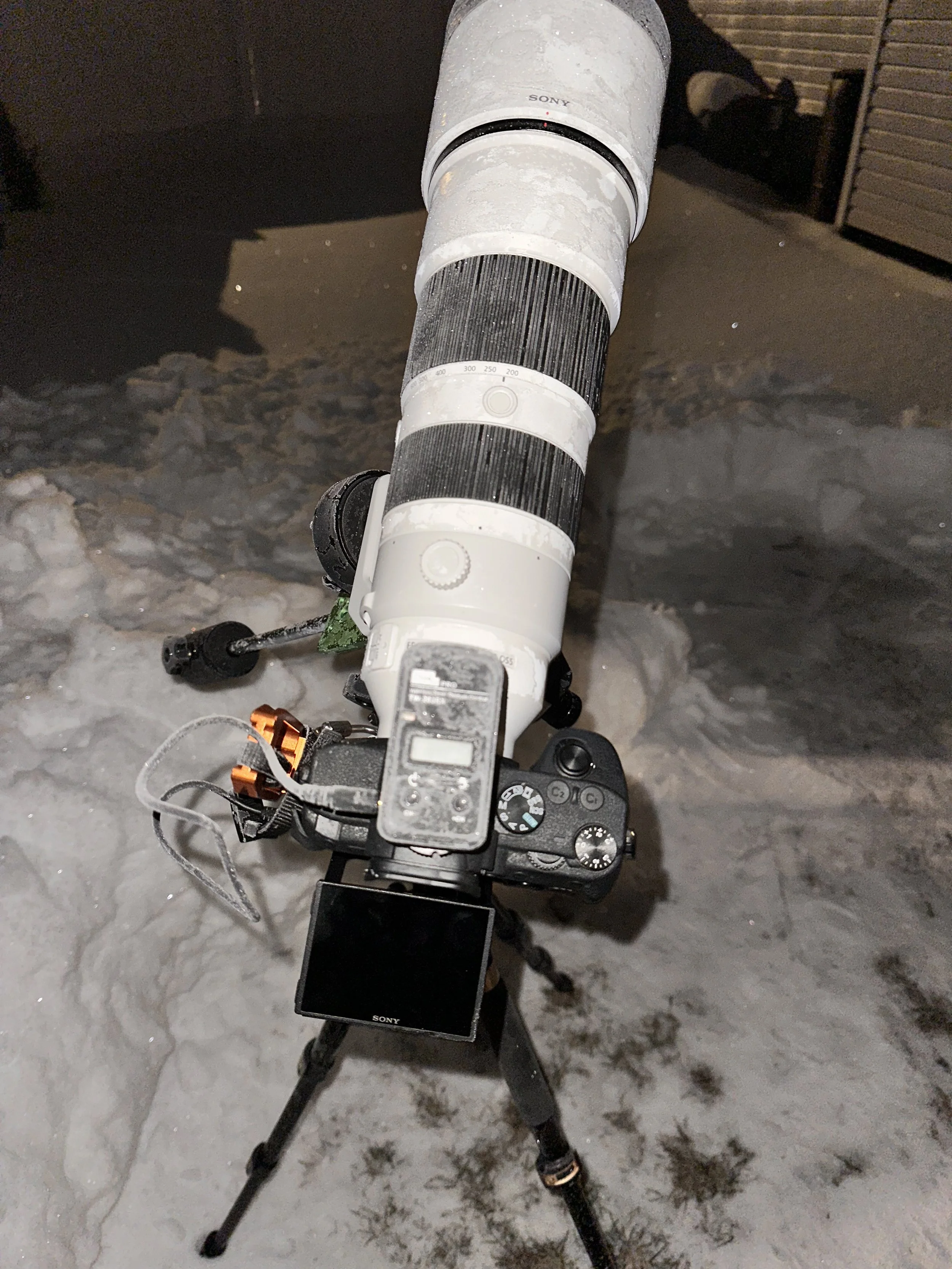 A camera mounted on a tripod, with a Sony camera body attached, a telescopic lens, and a smartphone mounted on top. The scene appears to be outdoors on an icy or snowy surface at night.