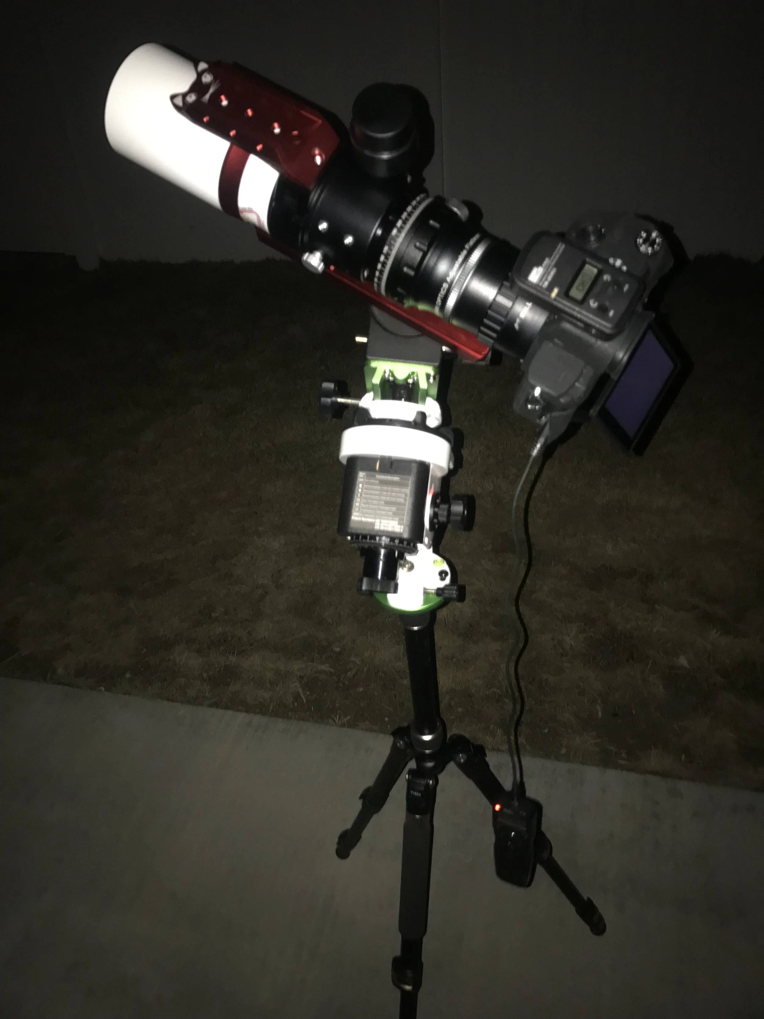 A telescope mounted on a tripod, pointed at the night sky, with a camera attached and a digital display screen.