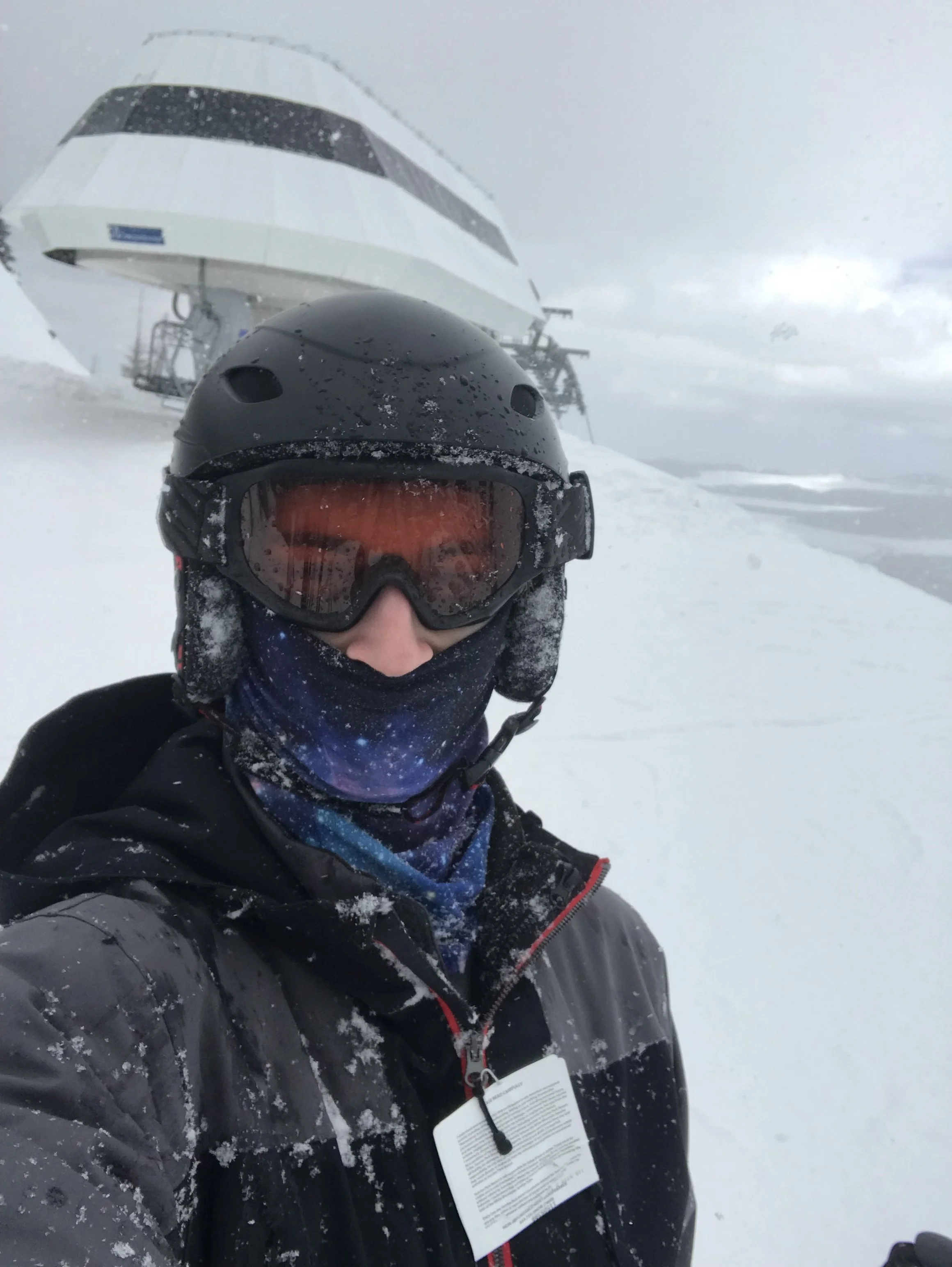 Person in black winter gear, including a helmet and goggles, taking a selfie in a snowy landscape with ski lift structures and cloudy sky in the background.