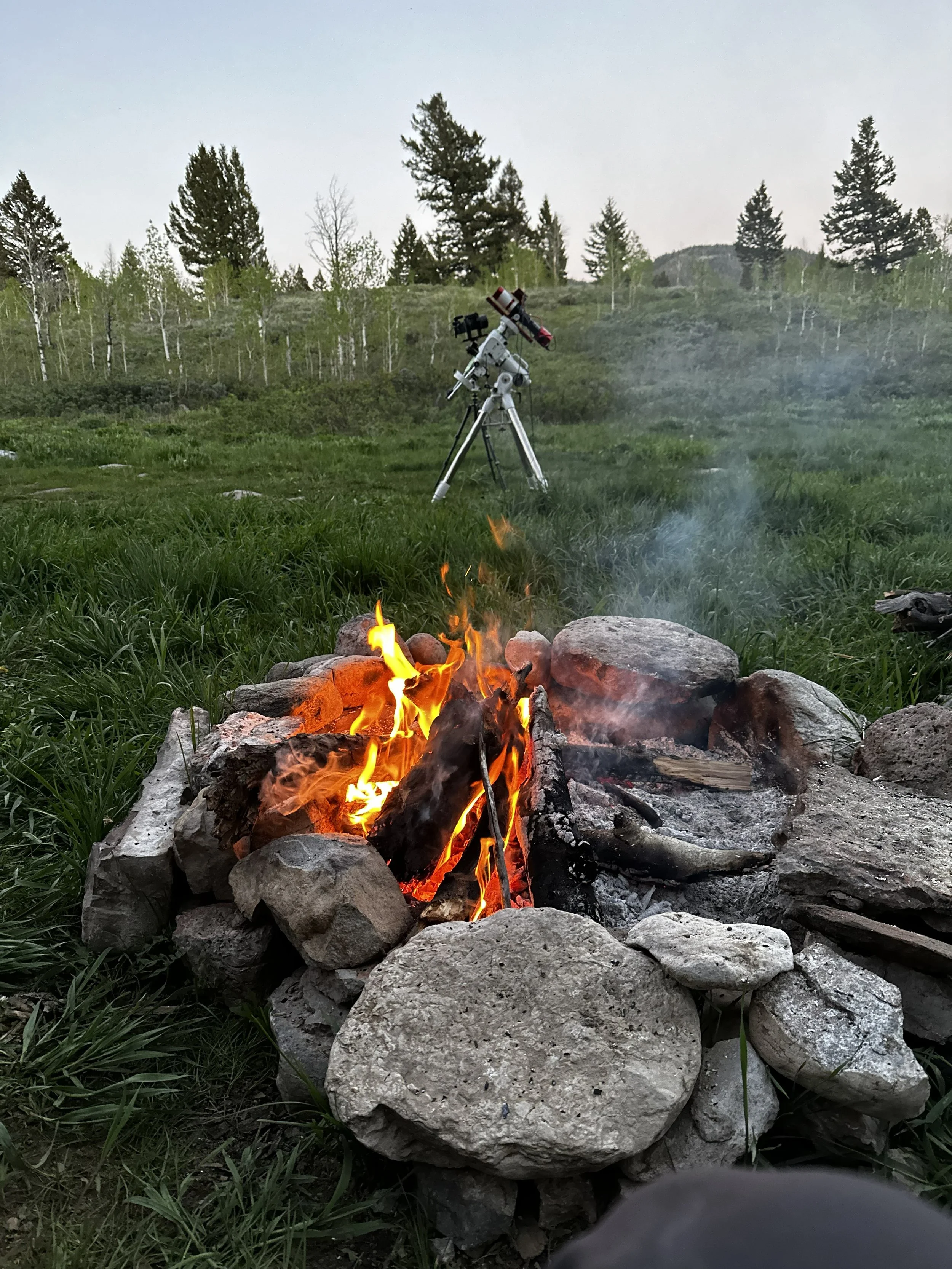 A campfire built with rocks and burning logs in a grassy outdoor area with trees and hills in the background. A telescope is set up on a tripod nearby, aimed at the sky.