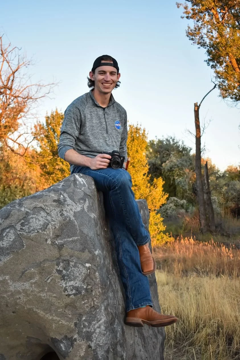 A young man sitting on a large rock outdoors during autumn, holding a camera, wearing a NASA sweatshirt, jeans, cowboy boots, and a backward baseball cap, smiling at the camera against a background of trees with orange and yellow leaves.