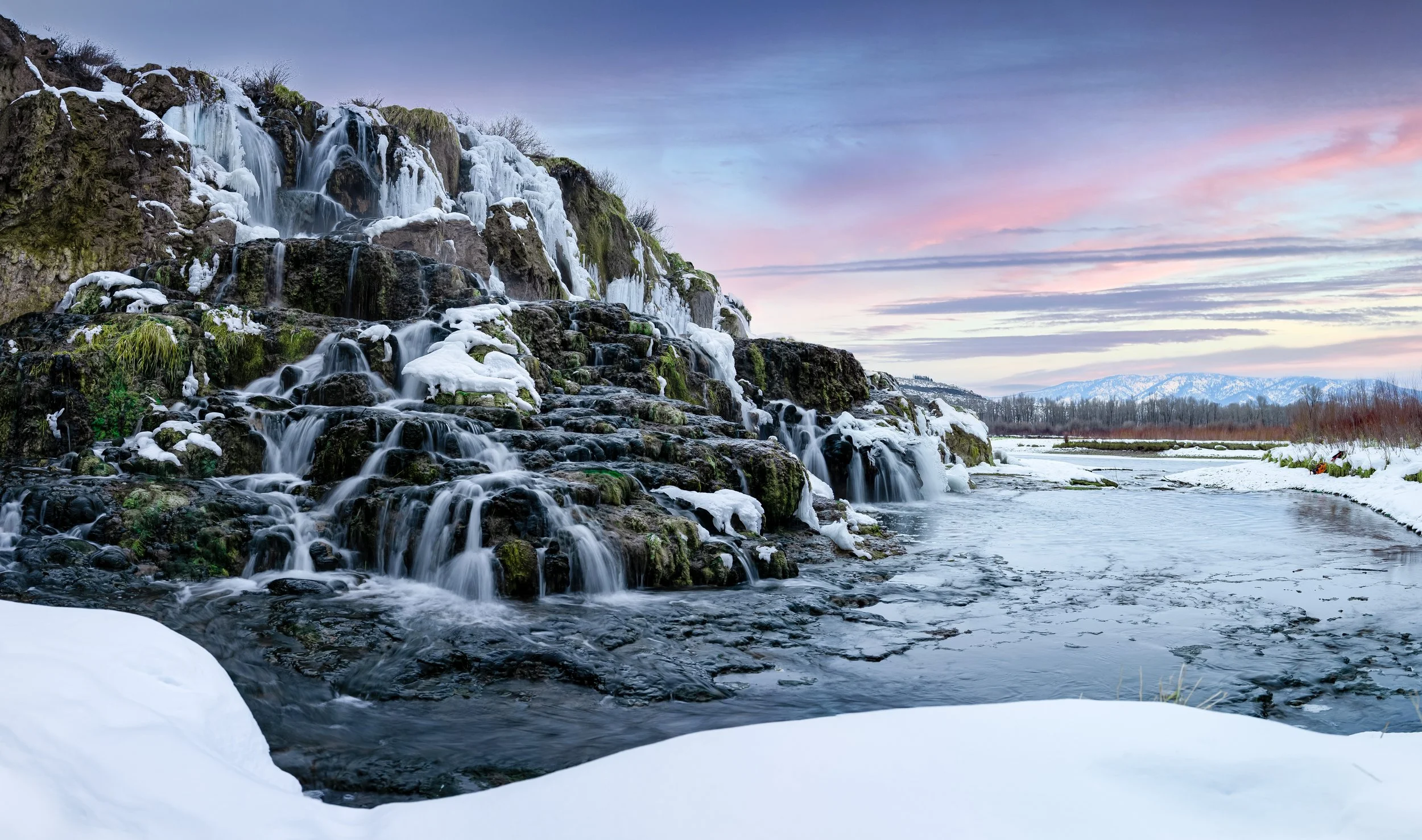 A cascading waterfall in a snowy landscape during winter with snow-covered rocks and trees, and a pale sky at dusk.
