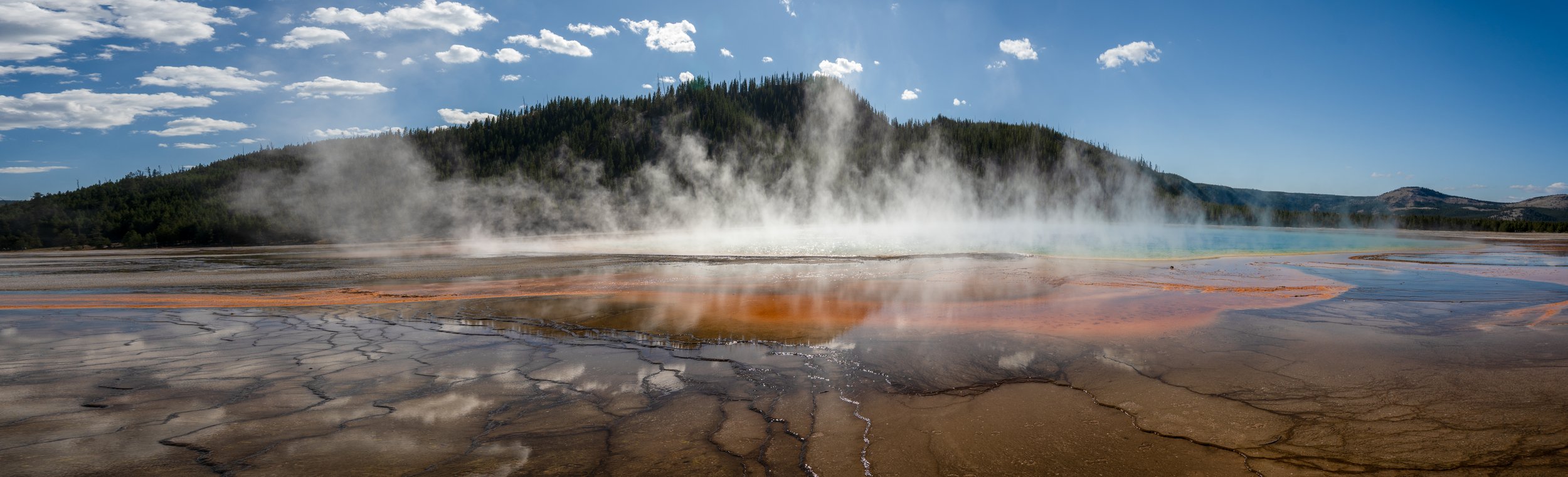 Hot springs with steam rising, colorful mineral deposits on the ground, mountains in the background, and a blue sky with clouds.
