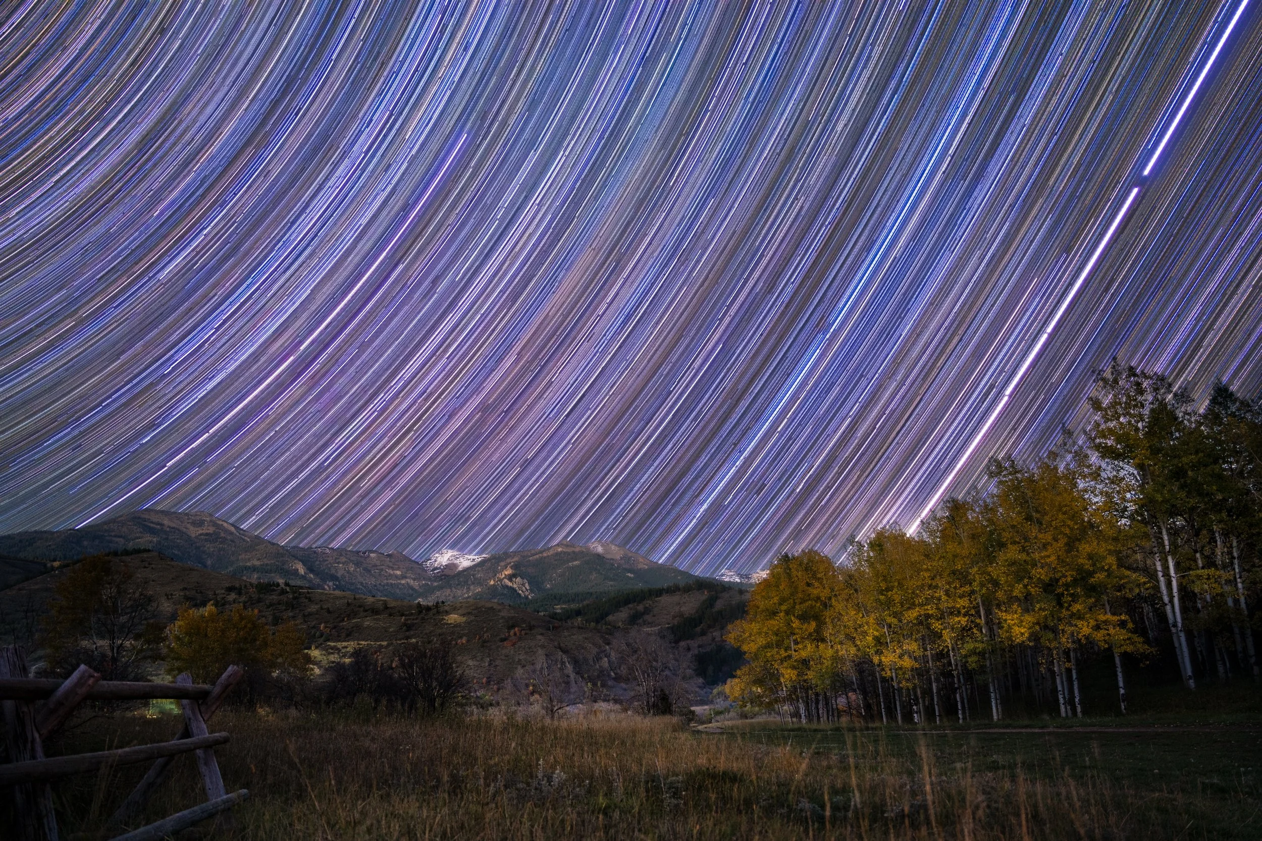 Long-exposure photograph of star trails arcing across the night sky over a rural landscape with trees, hills, and a wooden fence.