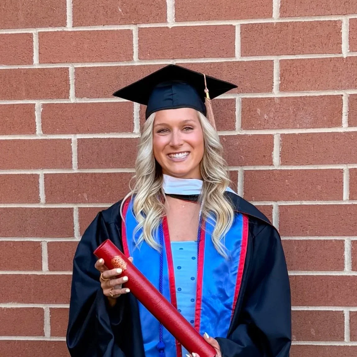 A young woman in a black graduation cap and gown, holding a diploma, standing in front of a brick wall, smiling.