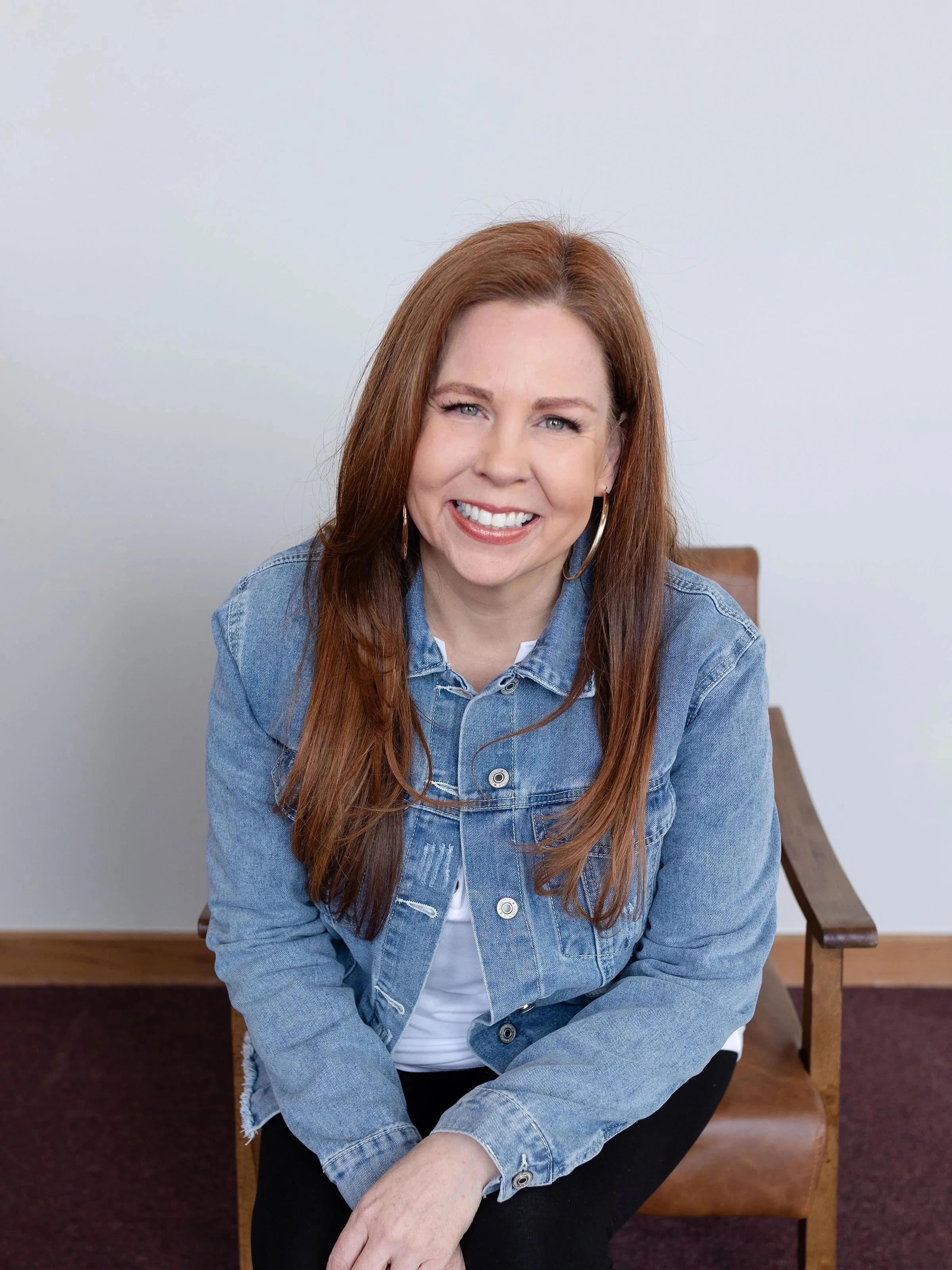A woman with long auburn hair smiling, wearing a blue denim jacket and black pants, sitting on a wooden chair.