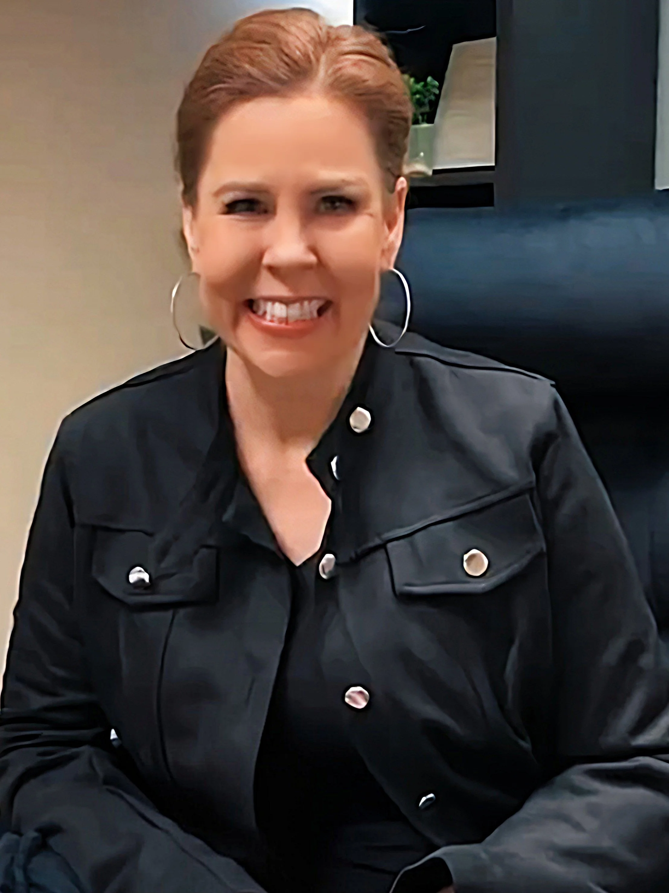 A woman with short reddish hair, smiling broadly, wearing large hoop earrings and a black jacket with silver buttons, sitting indoors.