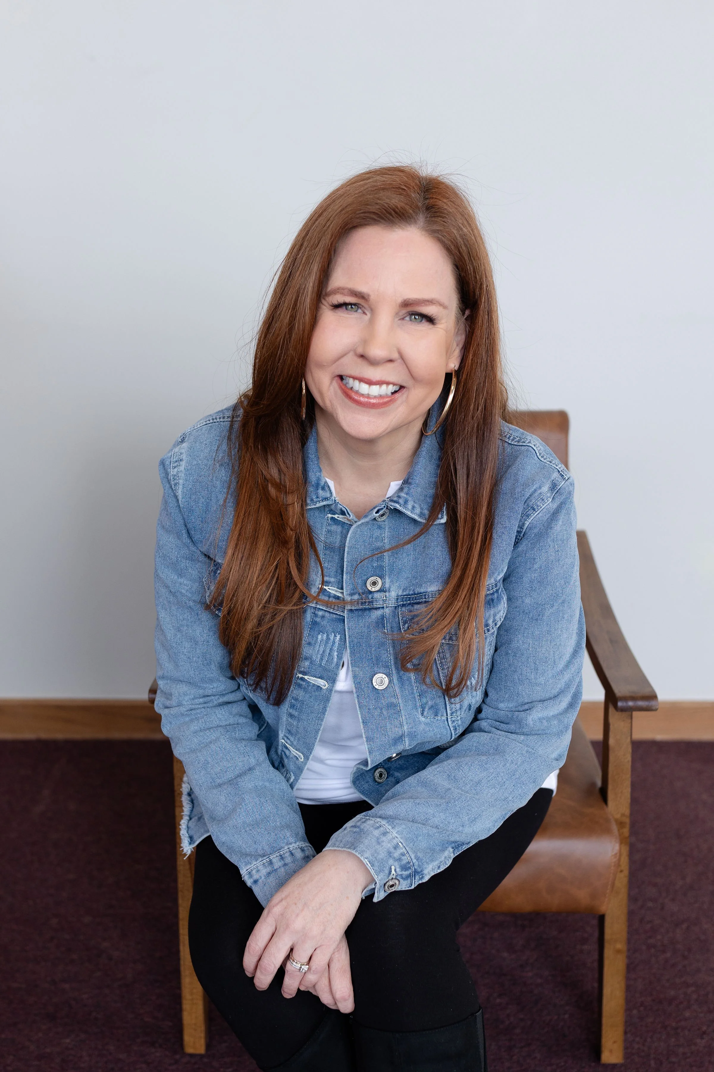 A woman with long reddish-brown hair, smiling, wearing a denim jacket over a white top, sitting on a wooden chair against a plain white wall.