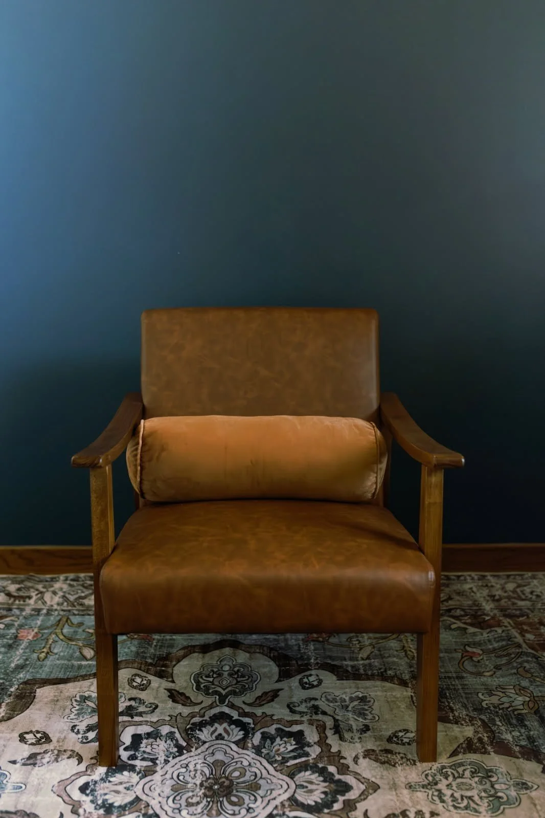 Vintage brown leather armchair with wooden armrests and a matching pillow, placed on a decorative rug against a blue wall.
