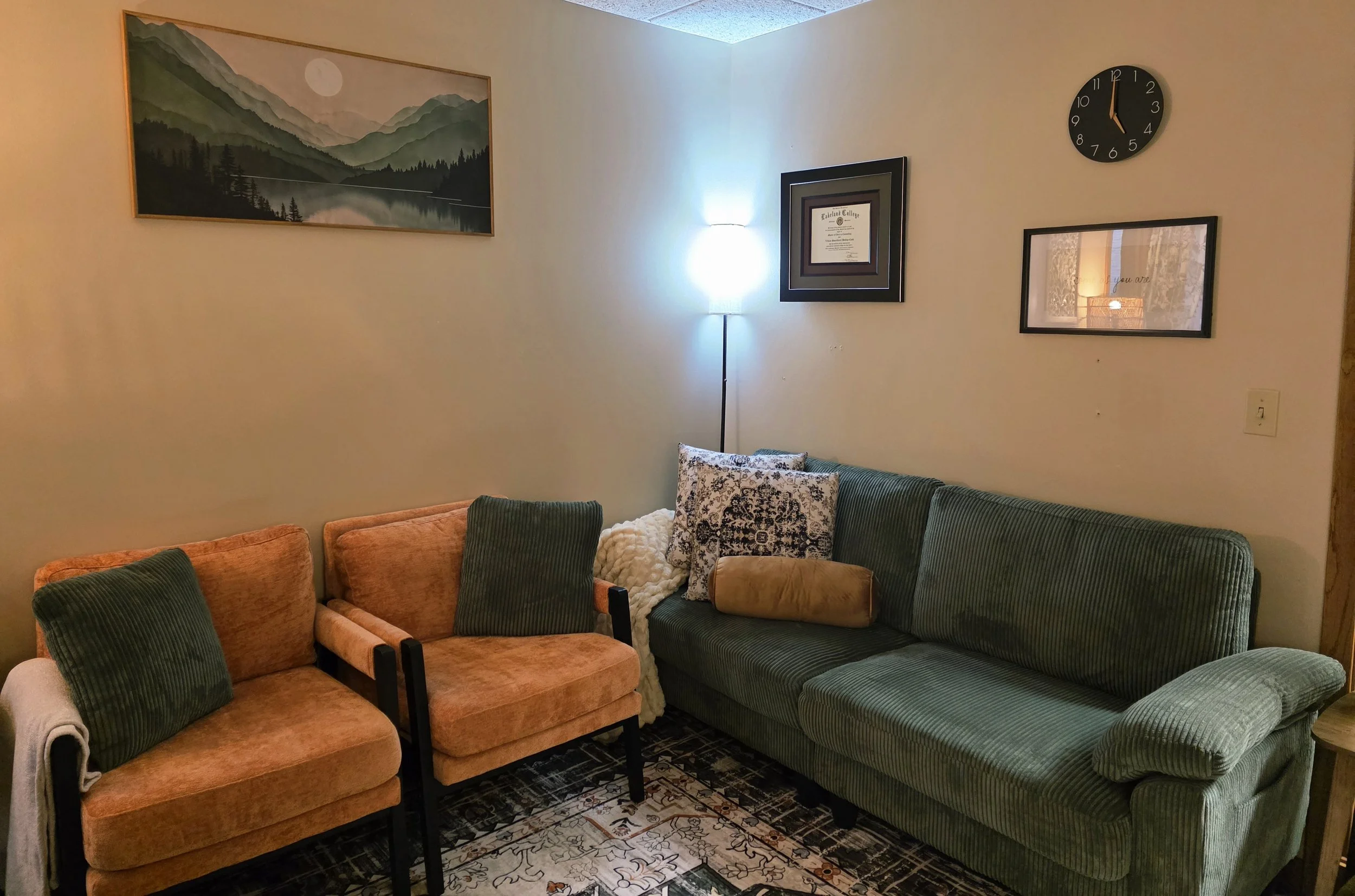A living room with a beige wall decorated with a landscape painting of mountains and a full moon, a framed diploma, and a circular black clock. There is a floor lamp in the corner. The room has a green and orange sofa with pillows, a patterned rug, and a window with lace curtains reflected in a wall mirror.