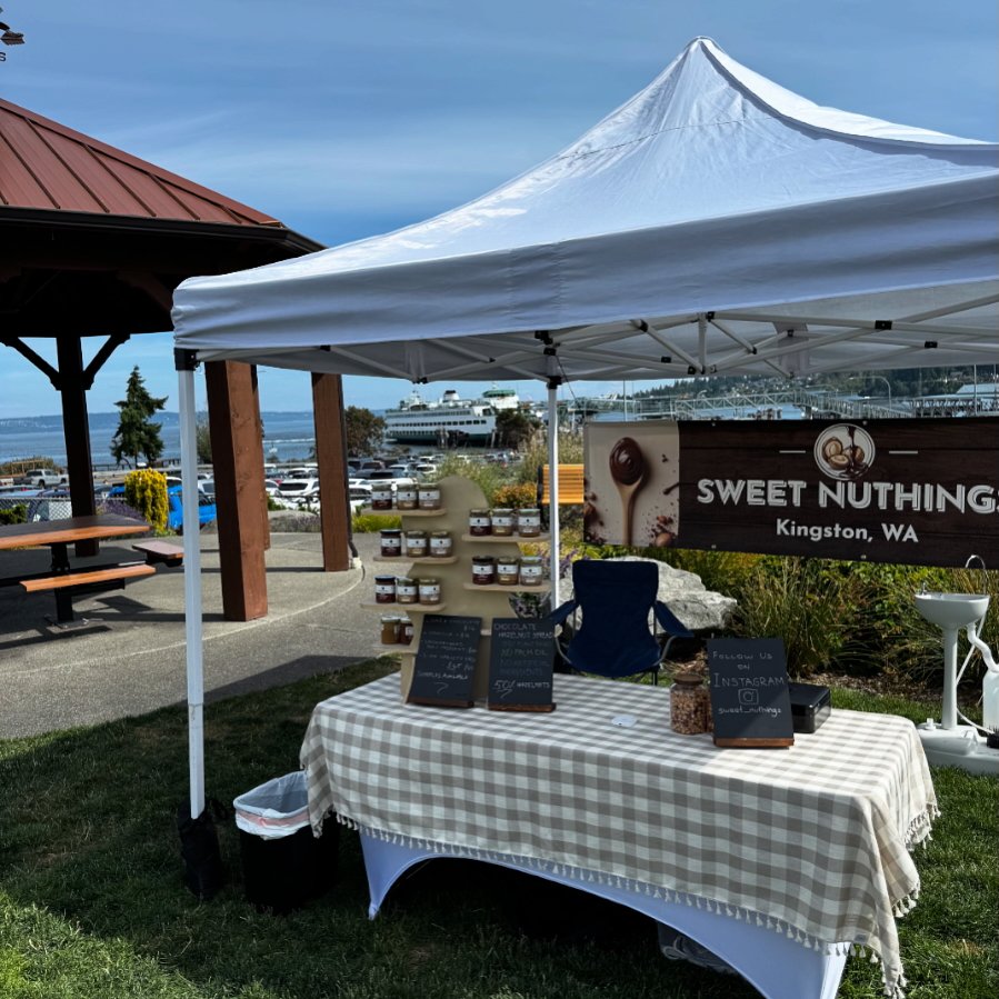 A small outdoor market stall with a white canopy tent, a checkered tablecloth, jars of honey or jam, and a sign that reads "SWEET NUTHING Kingston, WA". There is a parking lot with cars and a large ferry in the background near the waterfront.