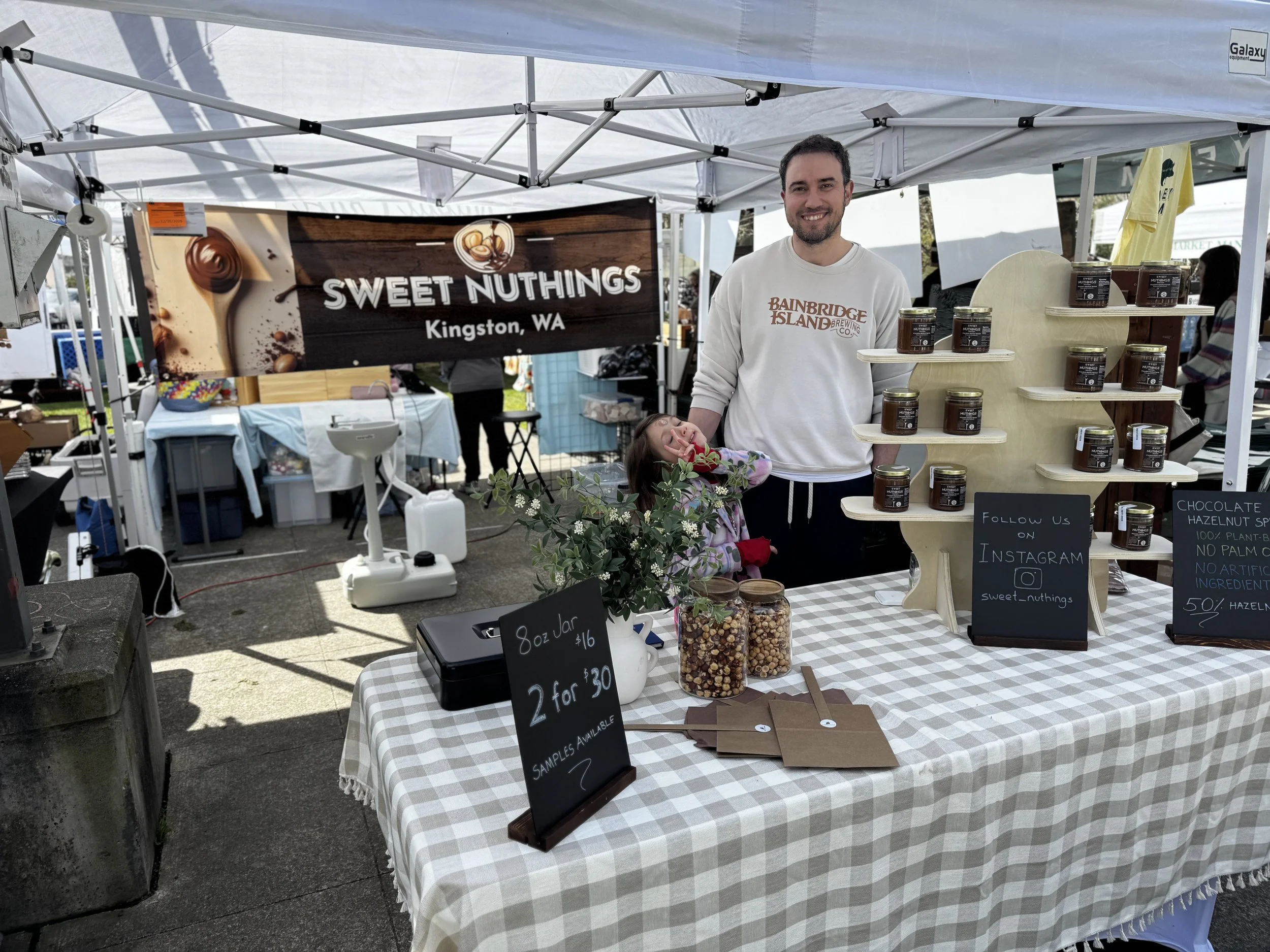 A man and a young girl standing at a vendor booth selling jars of nuts, with a sign reading 'Sweet Nuthings Kingston, WA'. The booth has a checkered tablecloth, jars of nuts, and signs displaying prices and social media information.