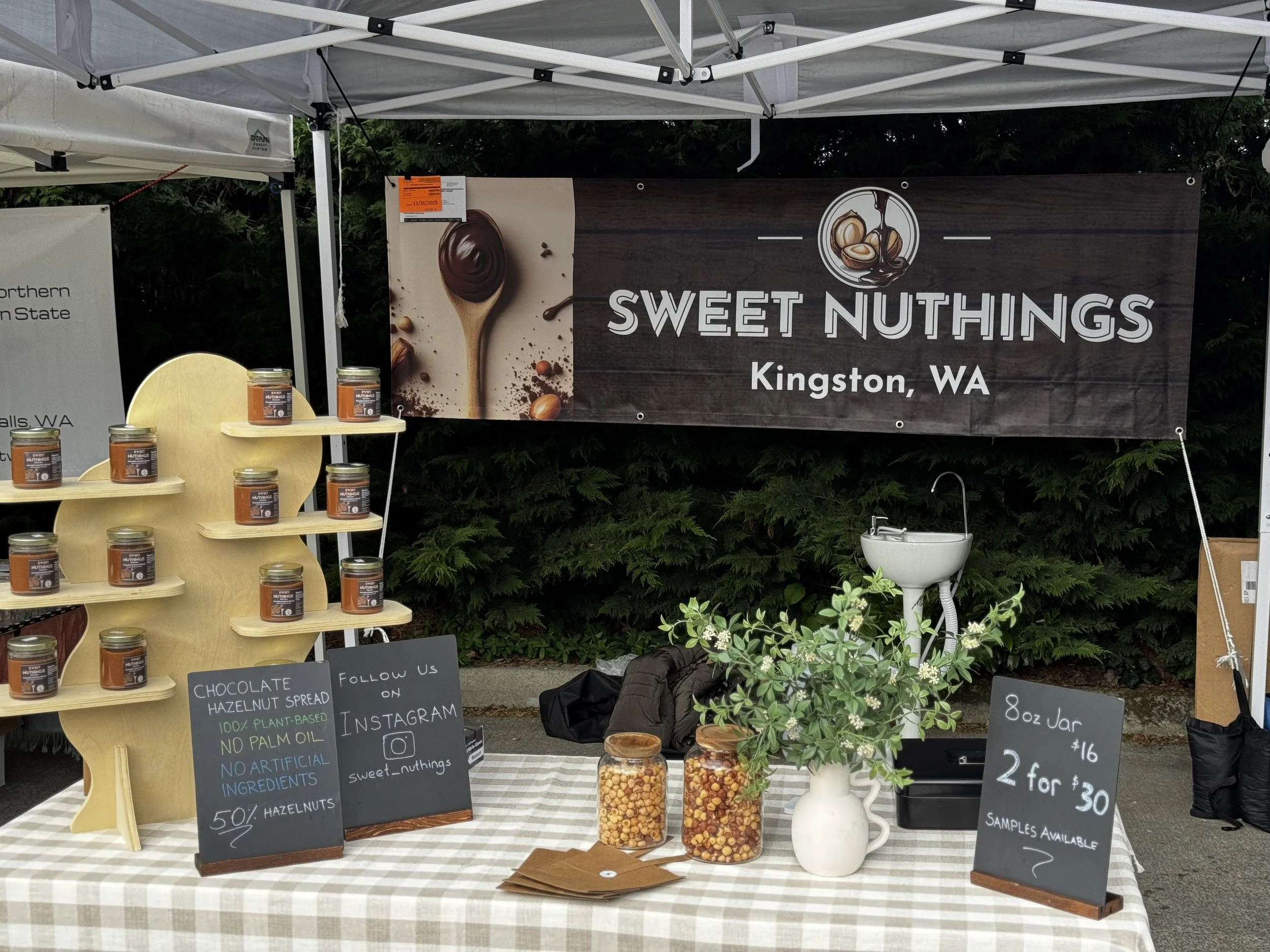 A market stall for Sweet Nuthings in Kingston, WA, selling chocolate hazelnut spread jars with a sign listing product details, jars of nuts, and sample information.