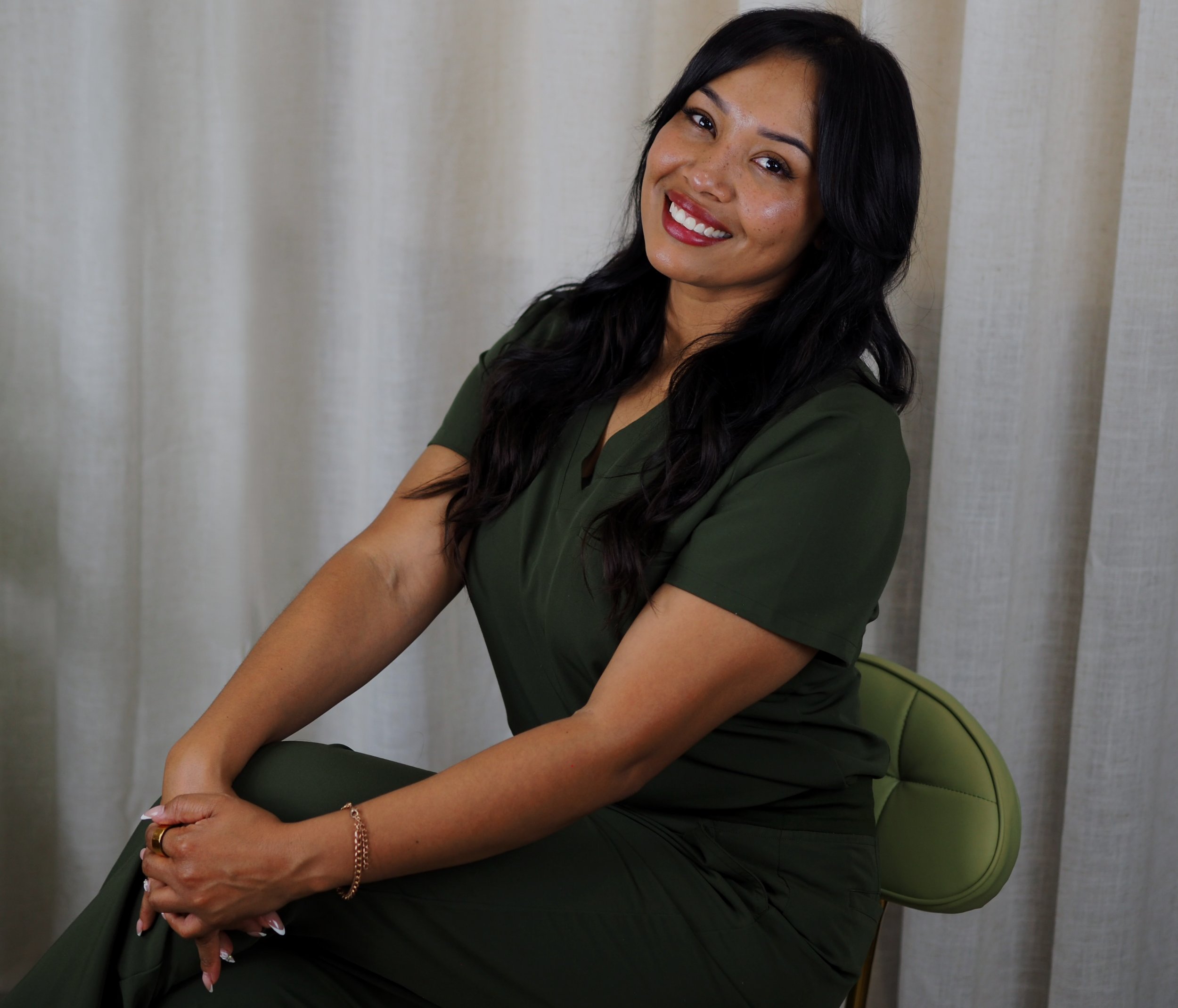 A woman with long black hair, wearing a dark green top, smiling with red lipstick, sitting on a green chair in front of a beige curtain.