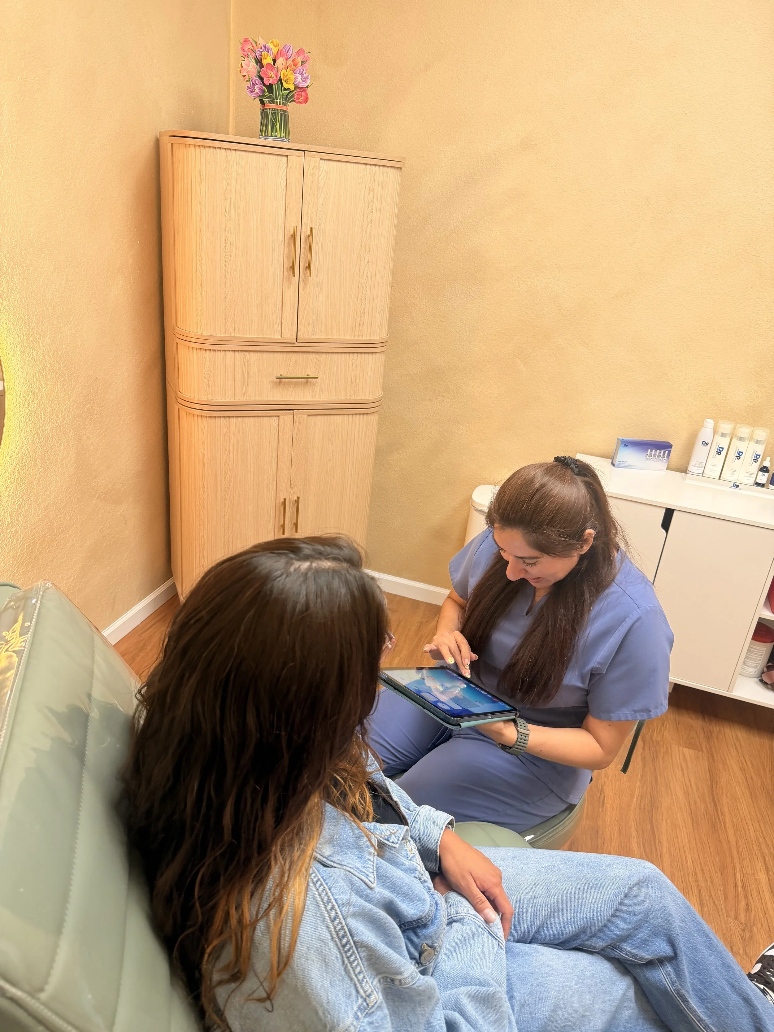 Three people sitting in a room, looking at a tablet. Two women in scrubs are engaged in a discussion, with a woman with long hair showing something on a tablet to another woman and a person with long hair wearing a denim jacket also seated nearby.