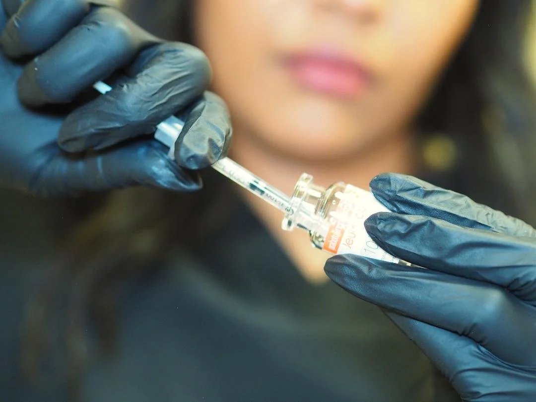 A person wearing black gloves holding a syringe and a small vial, preparing a vaccine or medication.