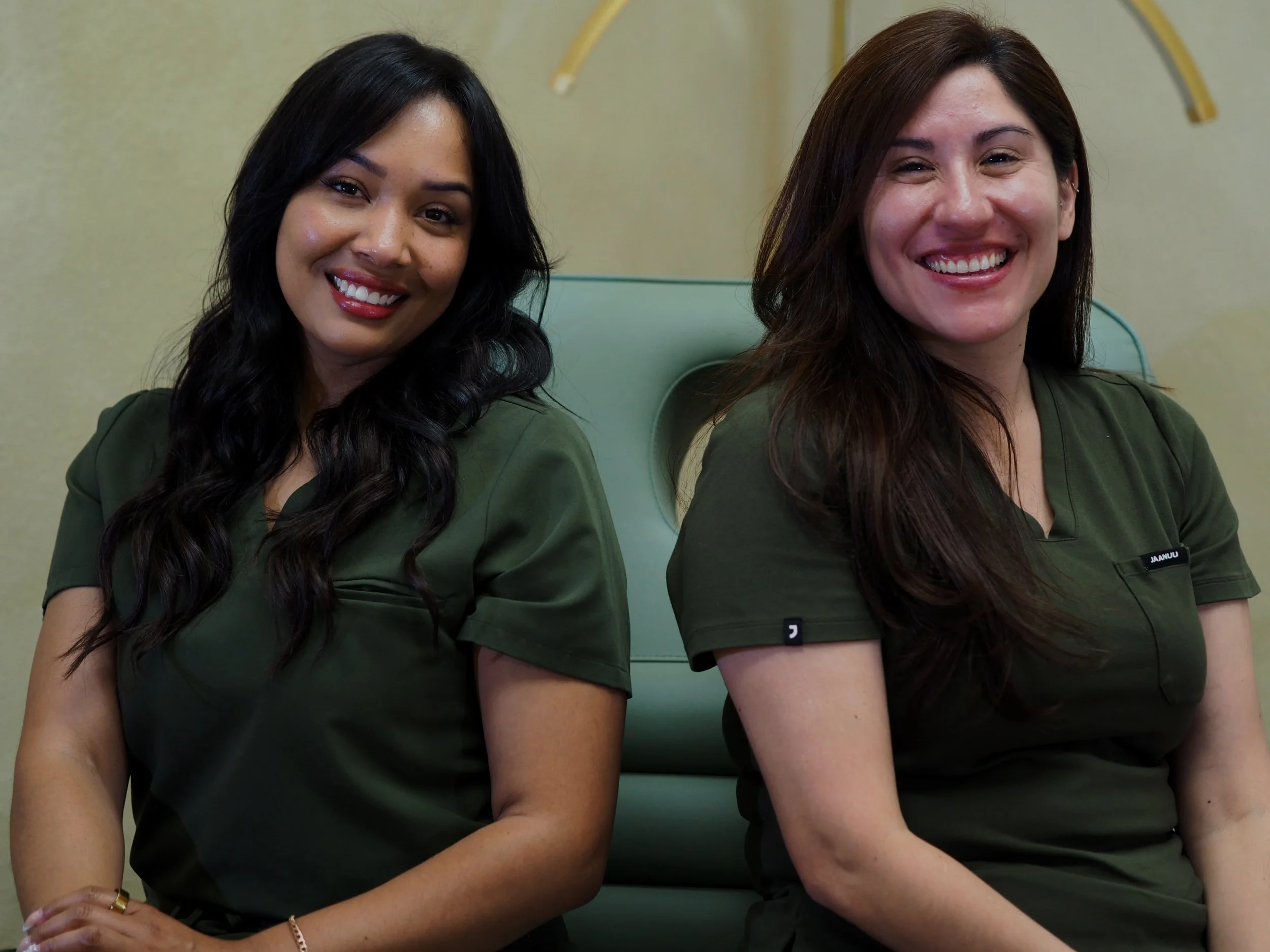Two women sitting close together, smiling at the camera. They are wearing green tops and are sitting in a room with a light-colored wall and a green chair.