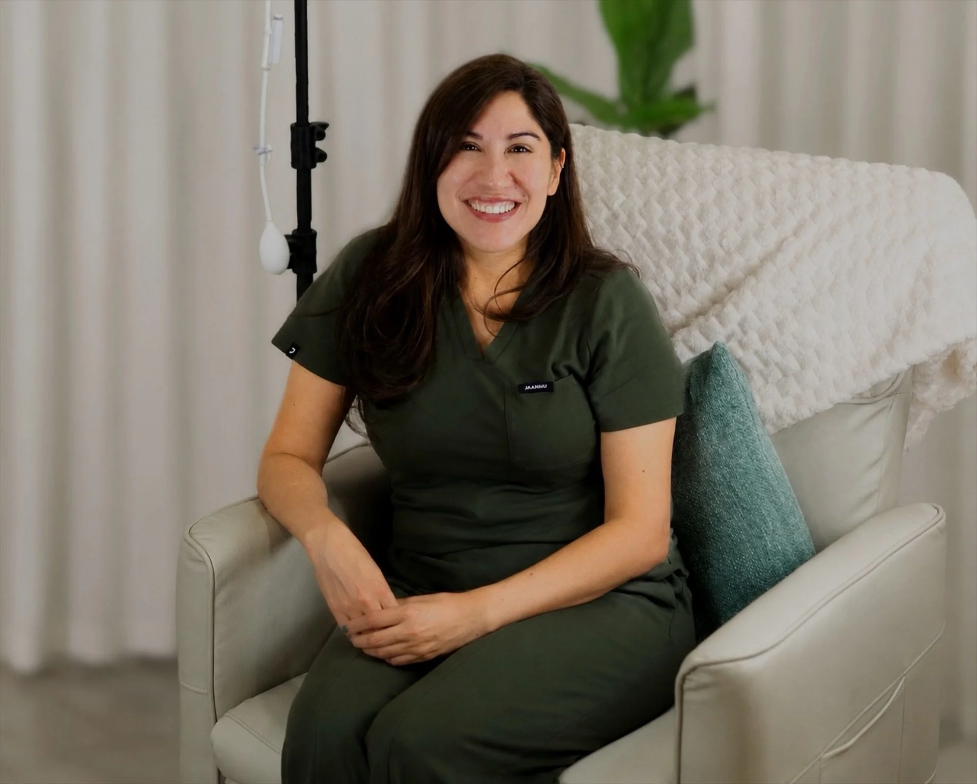 A woman with long brown hair smiling while sitting on a white armchair with a teal pillow in a room with white curtains and a large green leafy plant in the background.