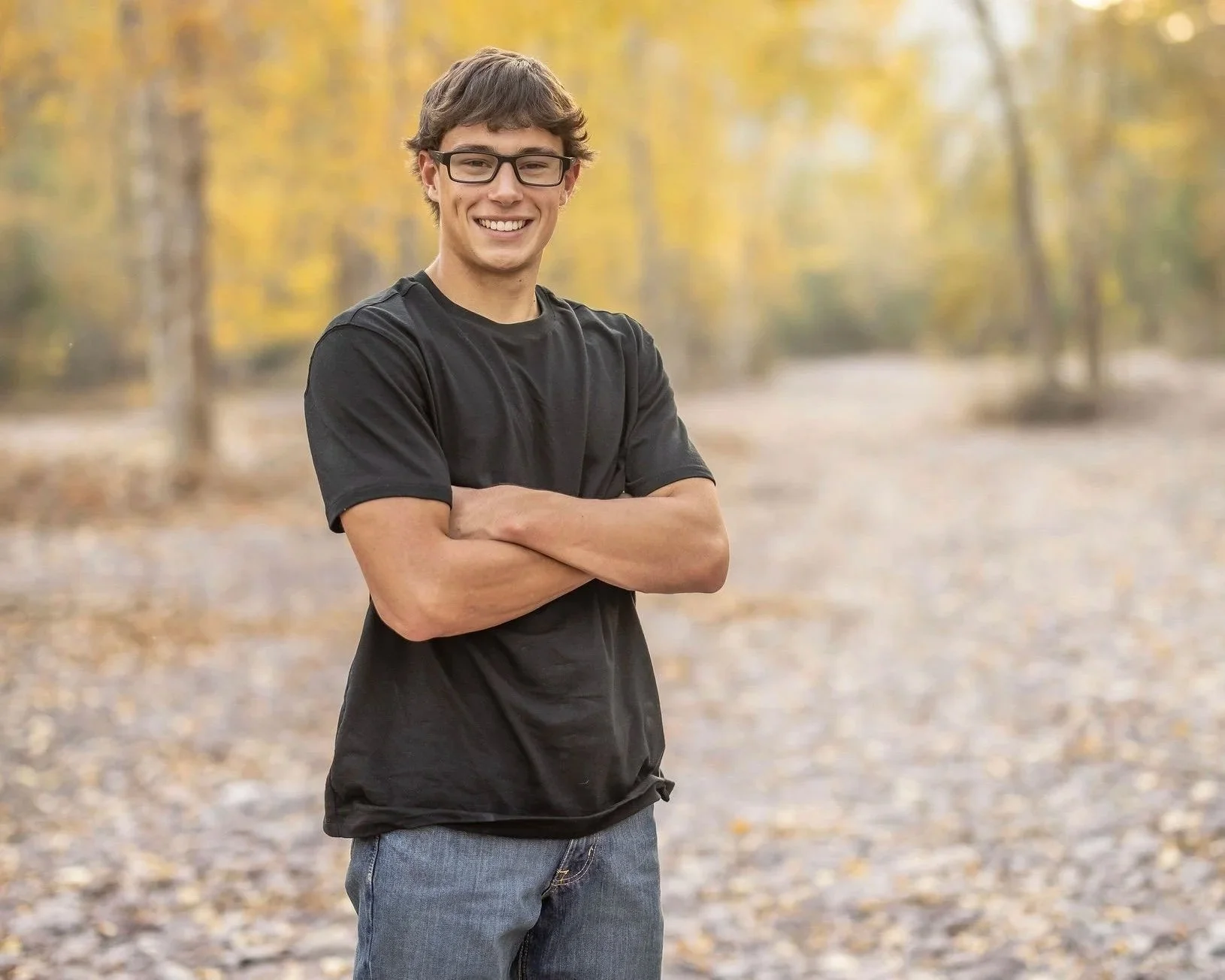 Young man with glasses smiling, standing outdoors with arms crossed, autumn trees in the background.