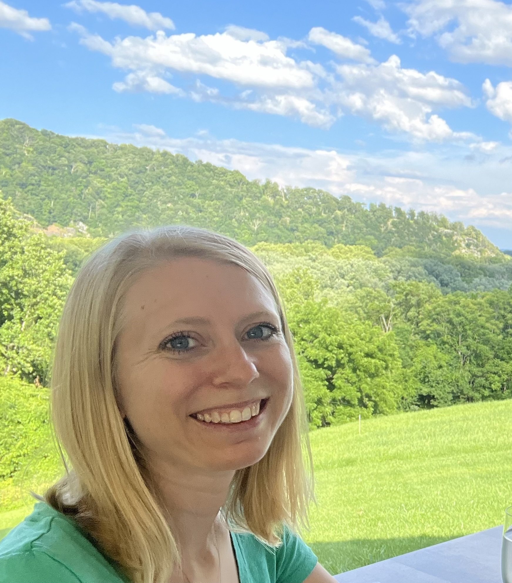 Smiling woman with blonde hair sitting outdoors with a background of green hills, trees, and a partly cloudy sky.