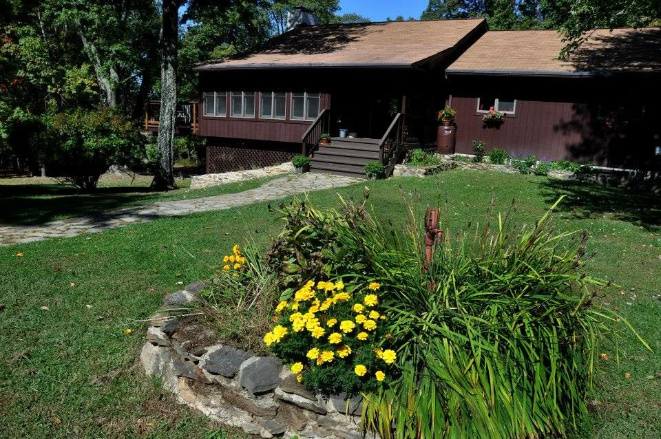 A brown house with a small porch, surrounded by greenery and a garden with yellow flowers in the front yard.