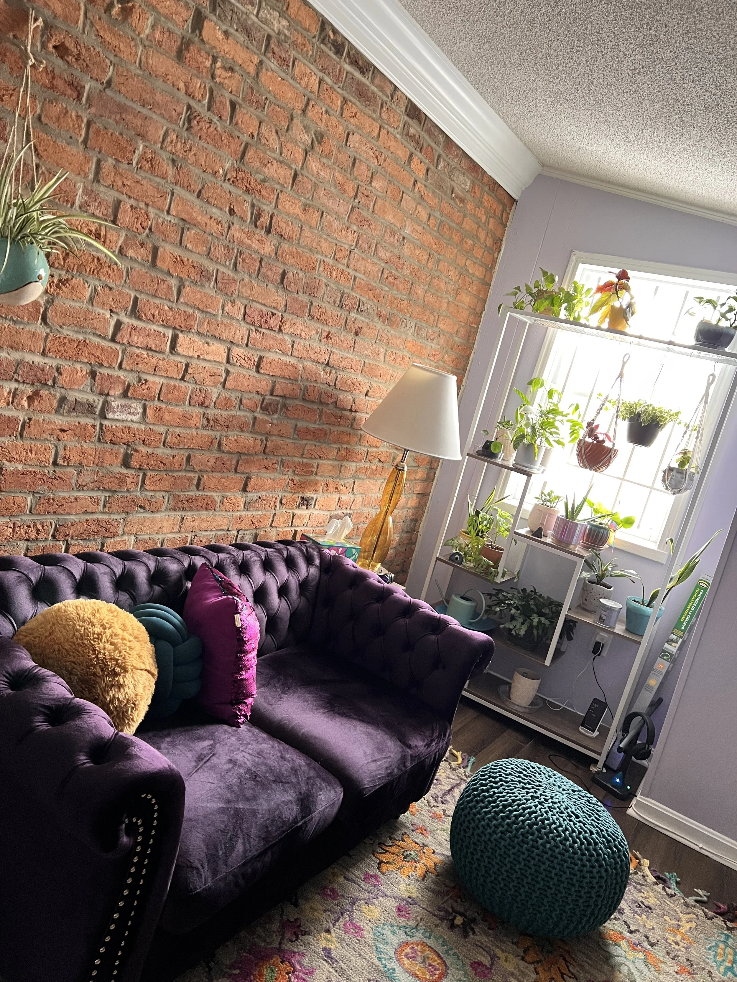 Living room with a purple velvet tufted sofa, pillows, a multicolored area rug, and a large green knitted pouf. A white shelving unit with plants and decor, a window with sunlight, and a floor lamp with a yellow base.