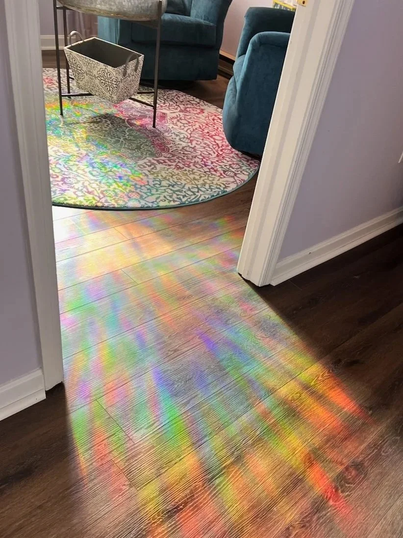 Colorful rainbow light reflections on wooden floor leading into a room with a colorful round rug, blue couches, and a black and white storage basket.