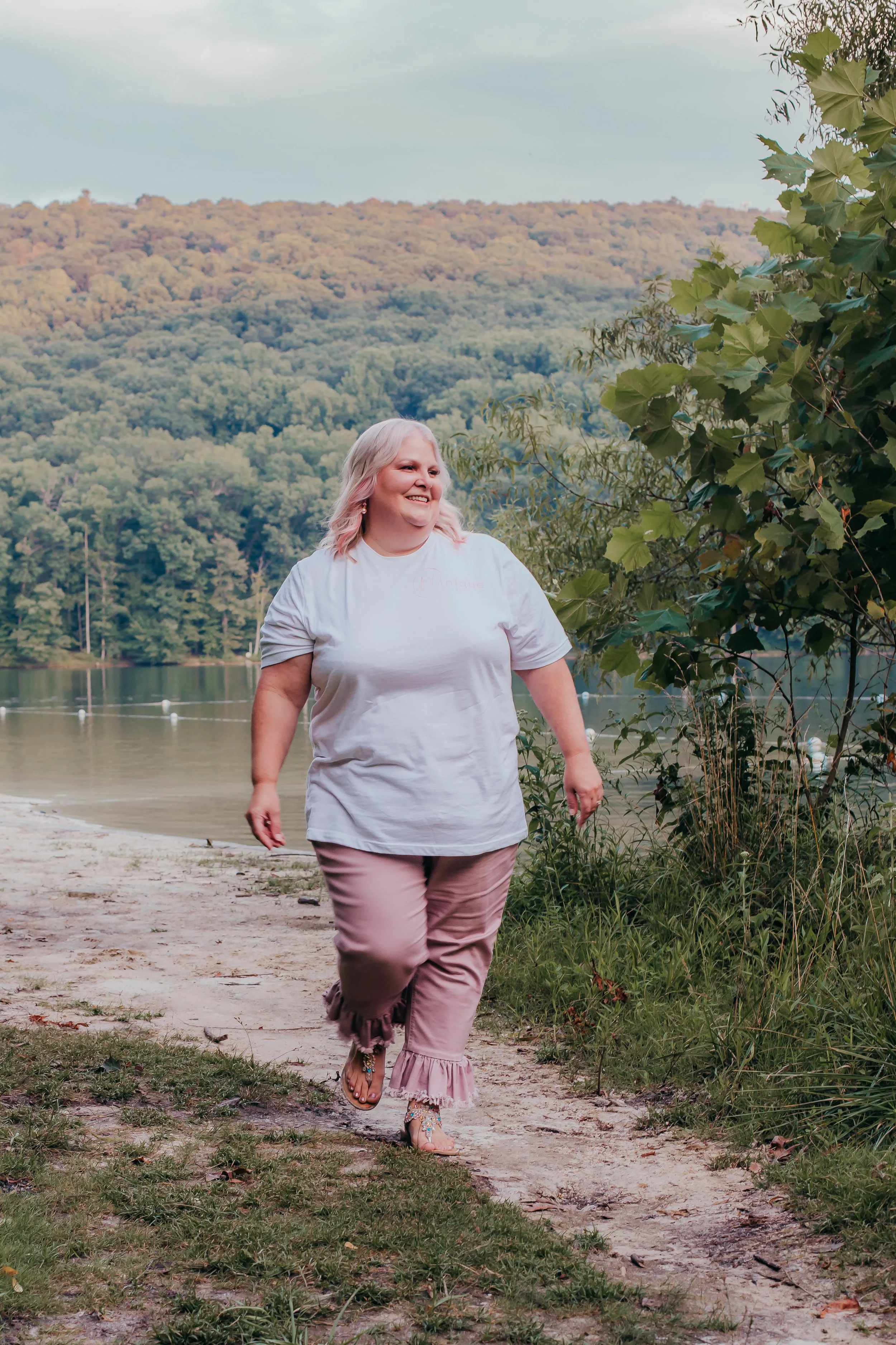 A woman with blonde hair walking on a dirt path near a lake, surrounded by trees and mountains in the background during daytime.