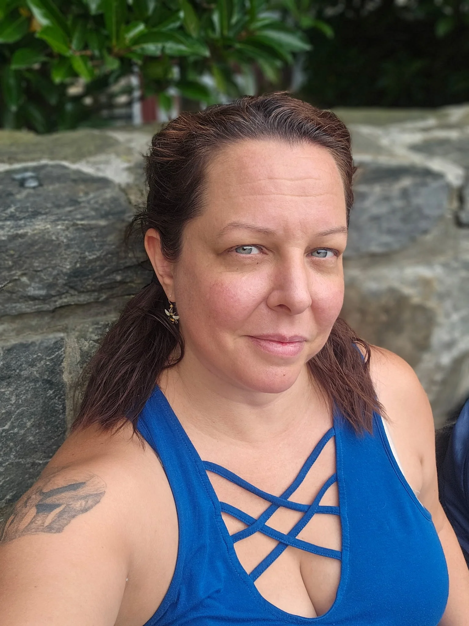A woman with light skin, brown hair, and blue eyes taking a selfie outdoors, wearing a blue top with crisscross straps and earrings, sitting in front of a stone wall with greenery in the background.