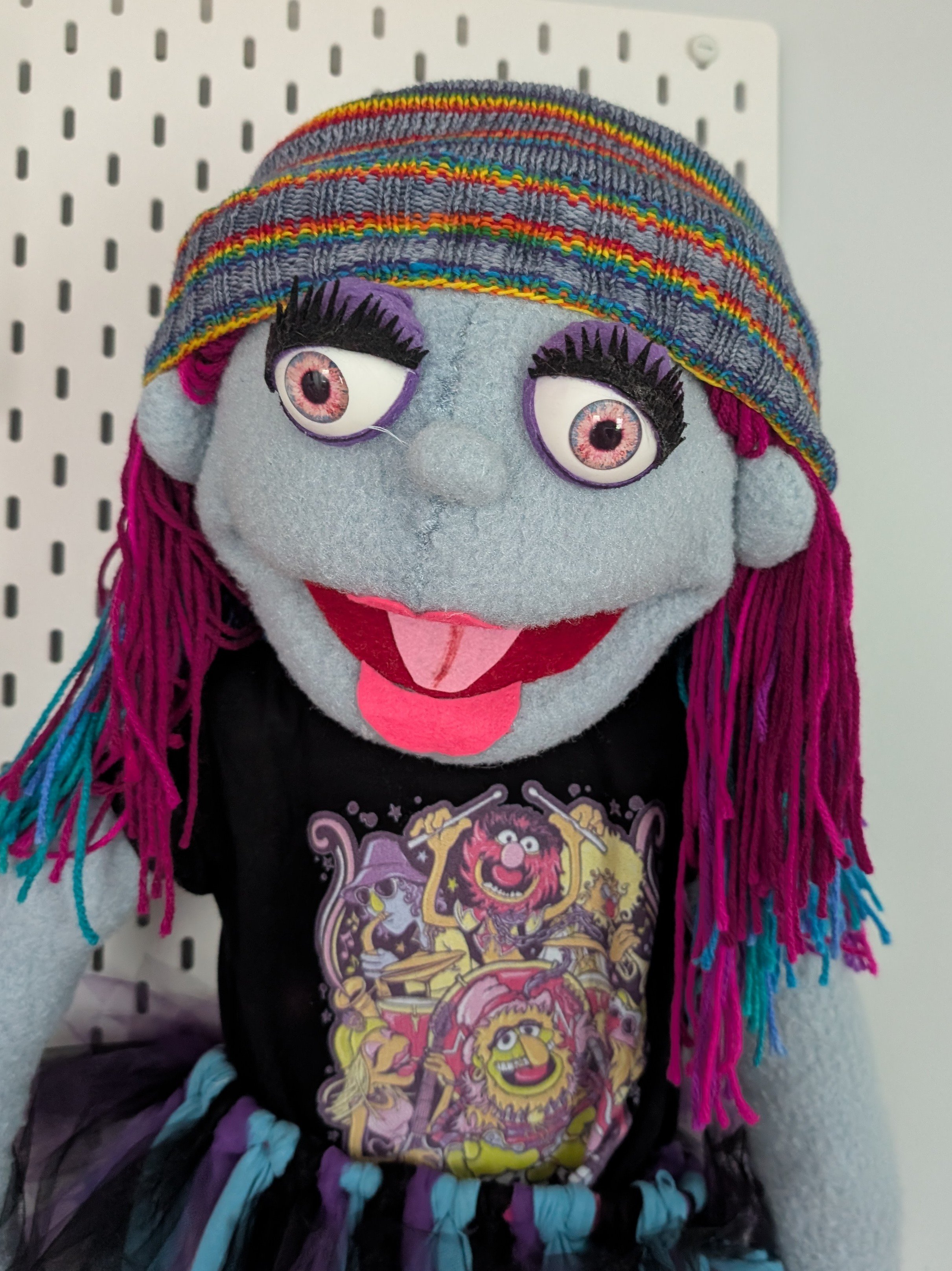 A puppet with a gray face, large eyes with pink irises, and long black eyelashes. It has pink and blue yarn hair, wears a colorful striped beanie, a black T-shirt with a cartoon band graphic, and a multicolored skirt.