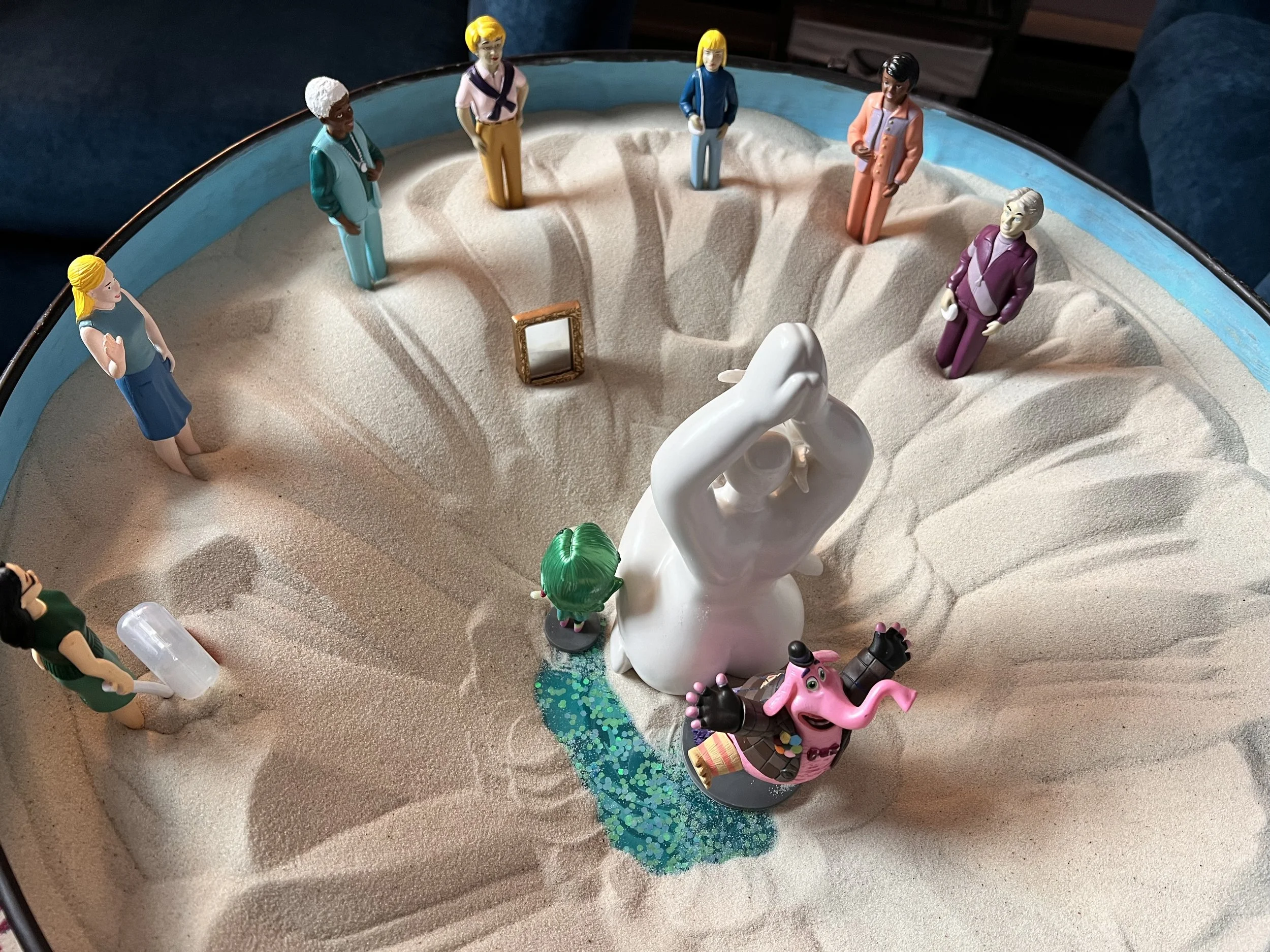 A sand tray with small figurines including women, men, and a cartoon pig, arranged around a small white sculpture with a tiny mirror and a green, glittery area.